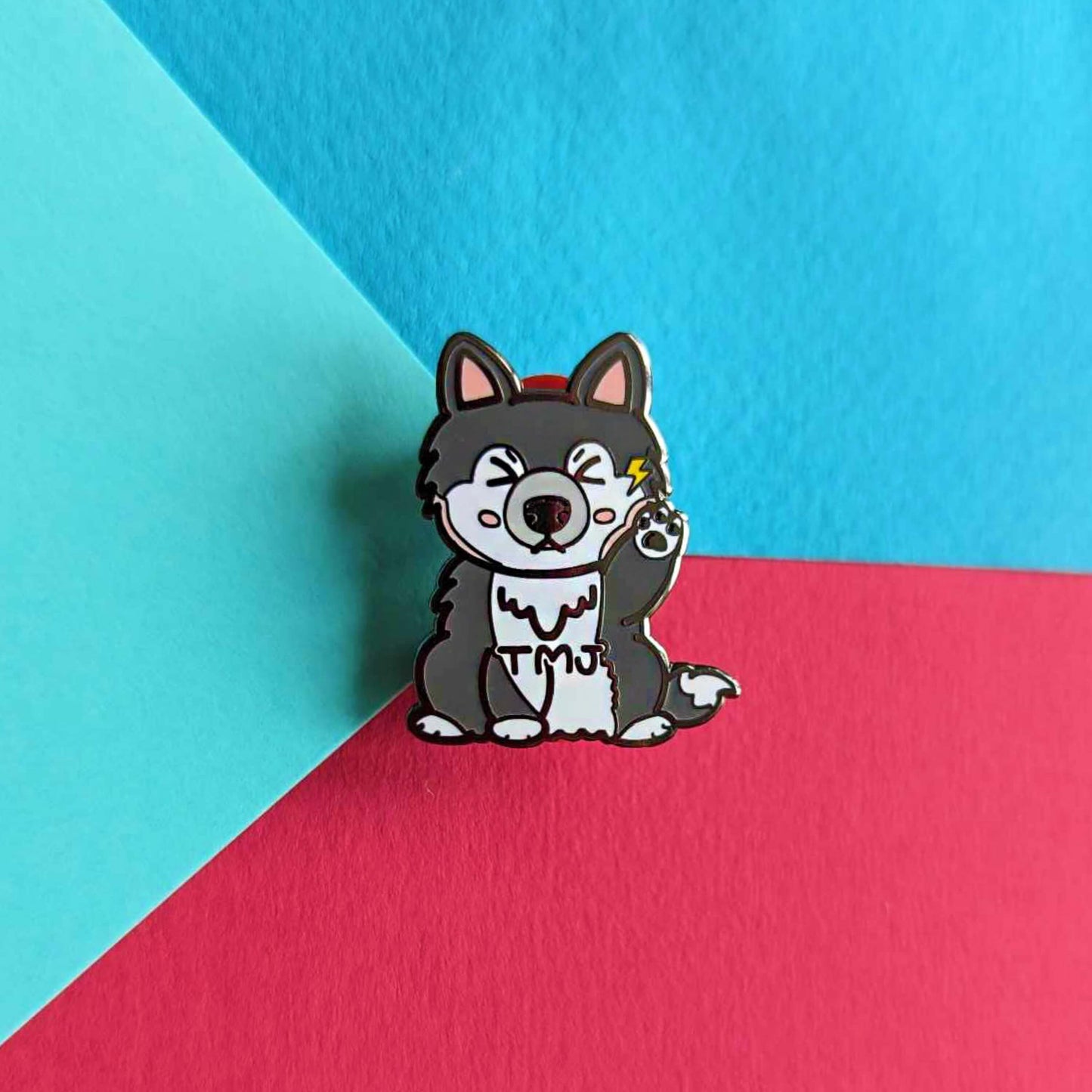  The Tempawromandibular Disorder Dog Enamel Pin - Temporomandibular Disorder TMJ on a blue and red background. The grey husky dog shaped enamel pin has a pained face with shut eyes, one paw up by its jaw with a yellow lightning bolt and black text across its chest reading 'TMJ'. The hand drawn design is raising awareness for Temporomandibular Disorder TMJ.