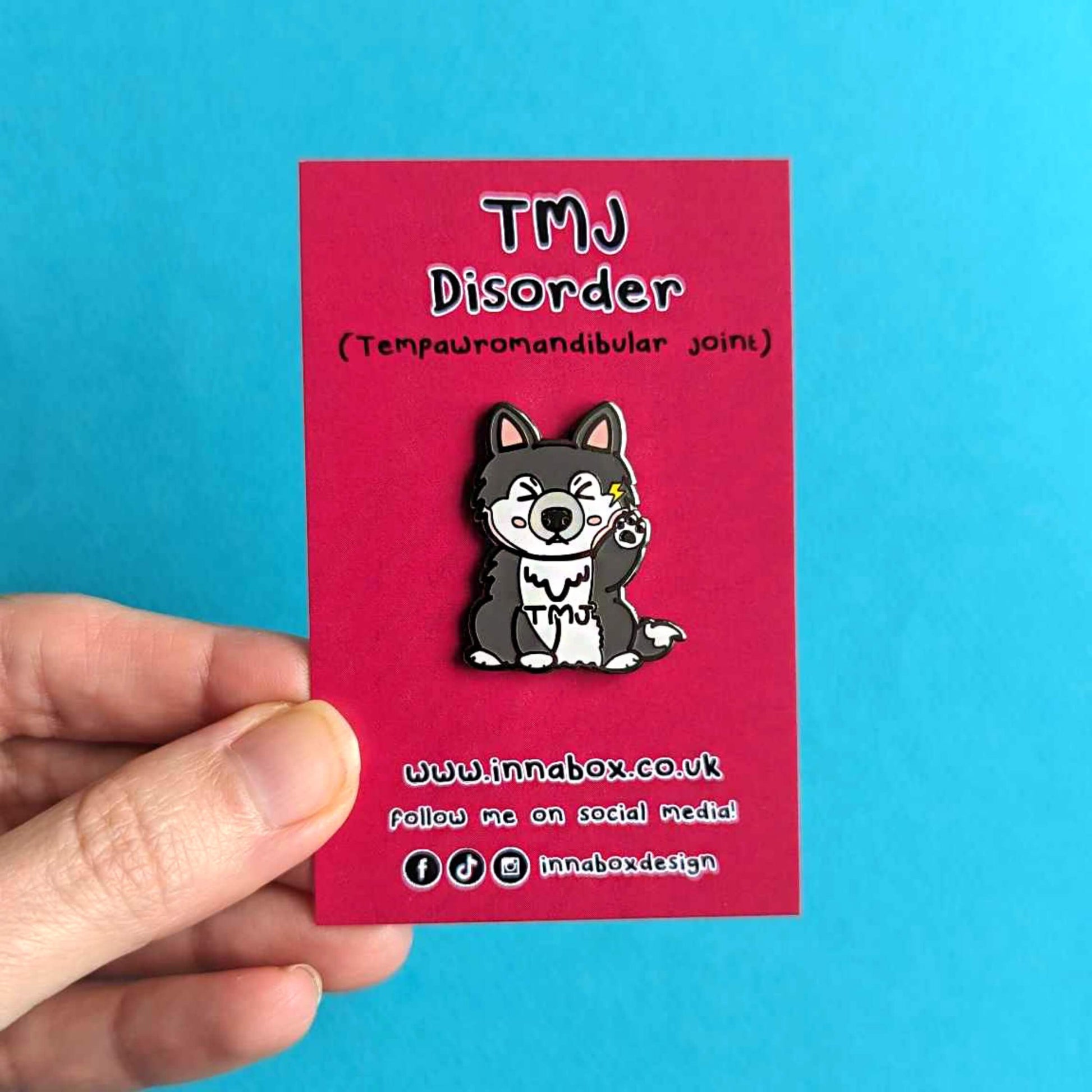 The Tempawromandibular Disorder Dog Enamel Pin - Temporomandibular Disorder TMJ on red backing card being held over a blue background. The grey husky dog shaped enamel pin has a pained face with shut eyes, one paw up by its jaw with a yellow lightning bolt and black text across its chest reading 'TMJ'. The hand drawn design is raising awareness for Temporomandibular Disorder TMJ.