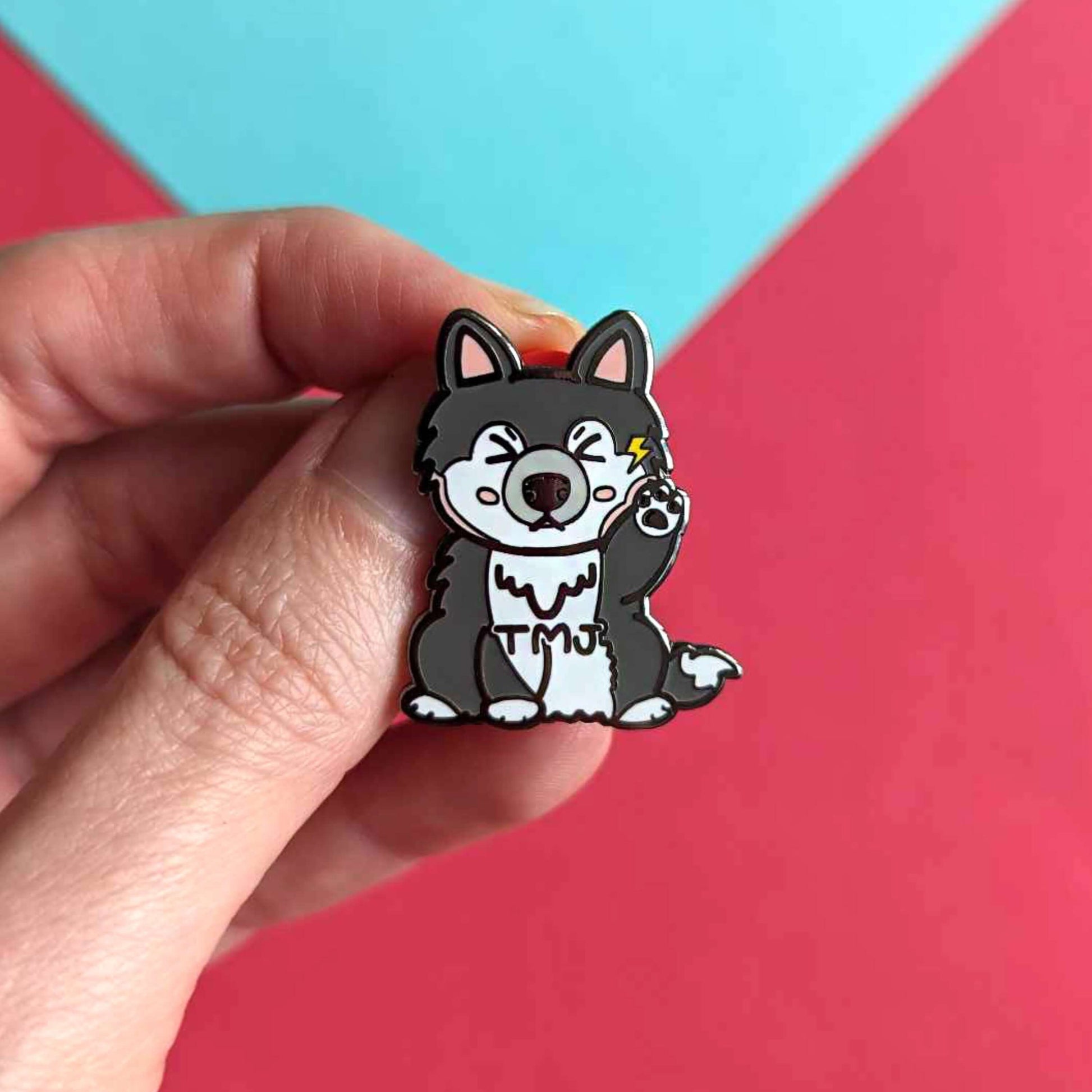 The Tempawromandibular Disorder Dog Enamel Pin - Temporomandibular Disorder TMJ being held over a blue and red background. The grey husky dog shaped enamel pin has a pained face with shut eyes, one paw up by its jaw with a yellow lightning bolt and black text across its chest reading 'TMJ'. The hand drawn design is raising awareness for Temporomandibular Disorder TMJ.