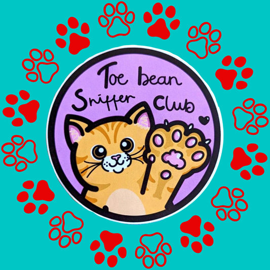 The Toe Bean Sniffer Club Sticker on a red and blue paw print background. The pastel pink circular sticker has a black outline and black text reading 'toe bean sniffer club' with a smiling orange cat raising a paw with a black heart.
