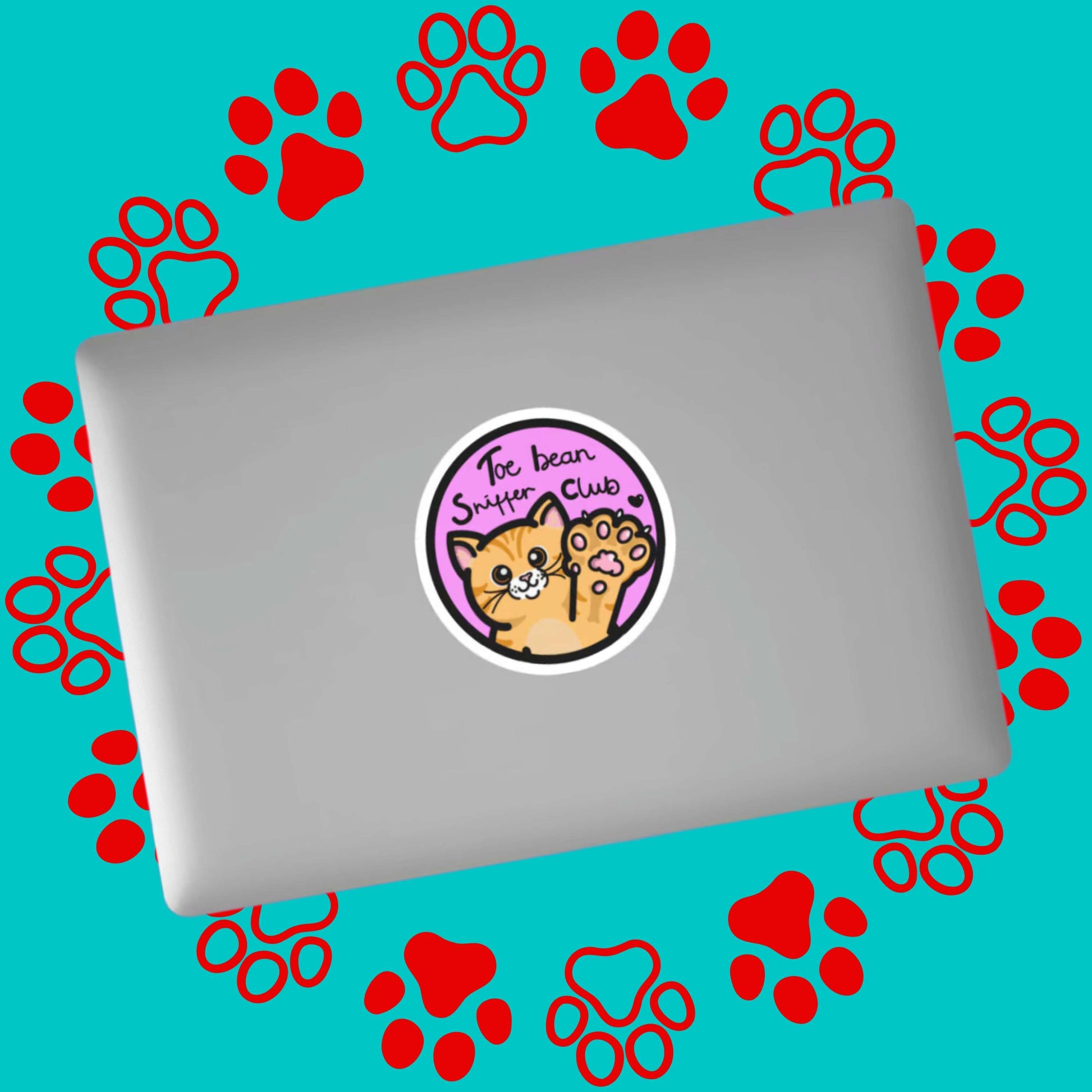 The Toe Bean Sniffer Club Sticker stuck on a silver laptop on a red and blue paw print background. The pastel pink circular sticker has a black outline and black text reading 'toe bean sniffer club' with a smiling orange cat raising a paw with a black heart.