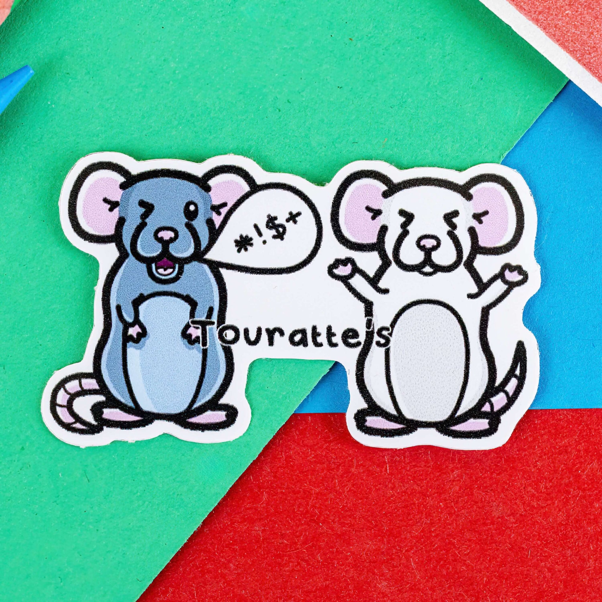The Touratte's Rat Sticker - Tourette's Syndrome on a red, blue and green background with colouring pencils and red stripe candy bag. The sticker features two rats, one grey rat on the left swearing in a speech bubble and one white rat with its eyes shut, across the middle in black text reads 'touratte's'. The hand drawn design is raising awareness for tourette's syndrome and tics.