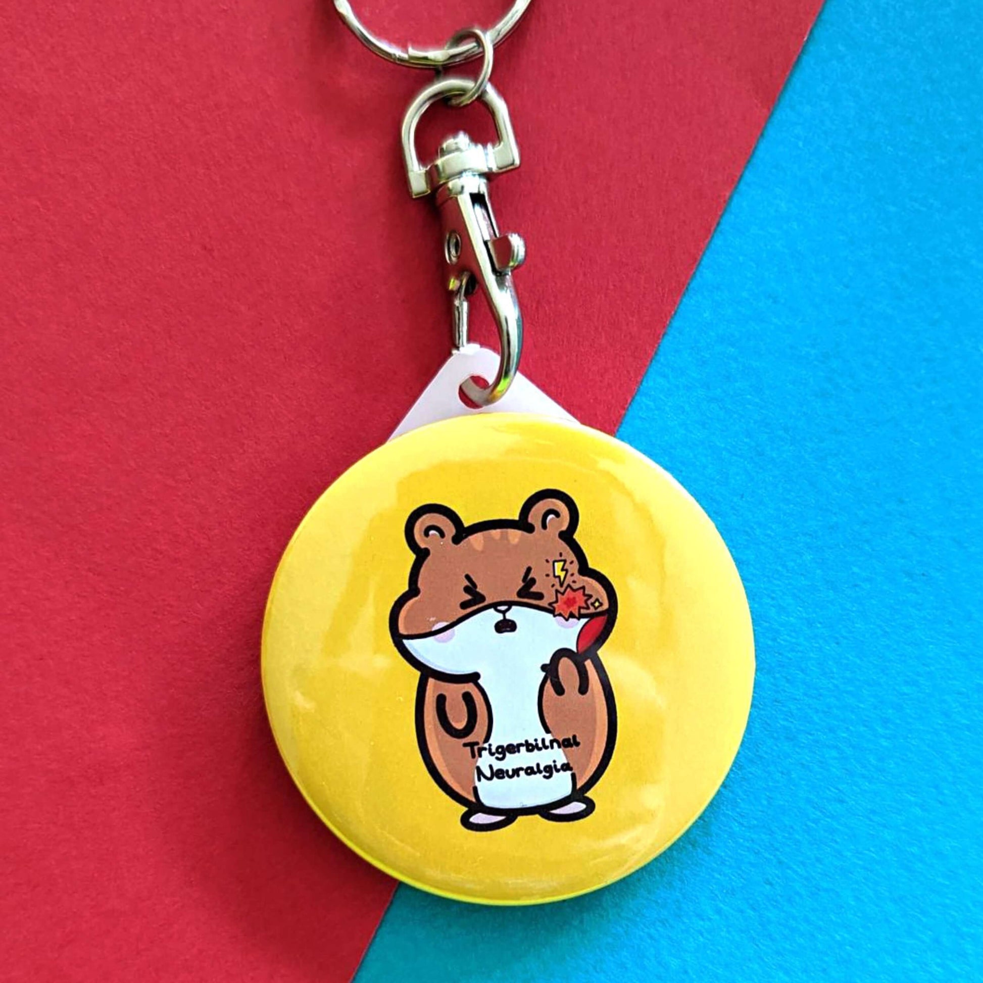 The Trigerbilnal Neuralgia Gerbil Keyring - Trigeminal Neuralgia TN on a red and blue background. The silver clip, yellow circular plastic keychain features a brown gerbil with a pained expression clutching its jaw that has a red explosion and yellow lightning bolt, across its middle in black reads 'Trigerbilnal Neuralgia'. The hand drawn design is raising awareness for Trigeminal Neuralgia TN.