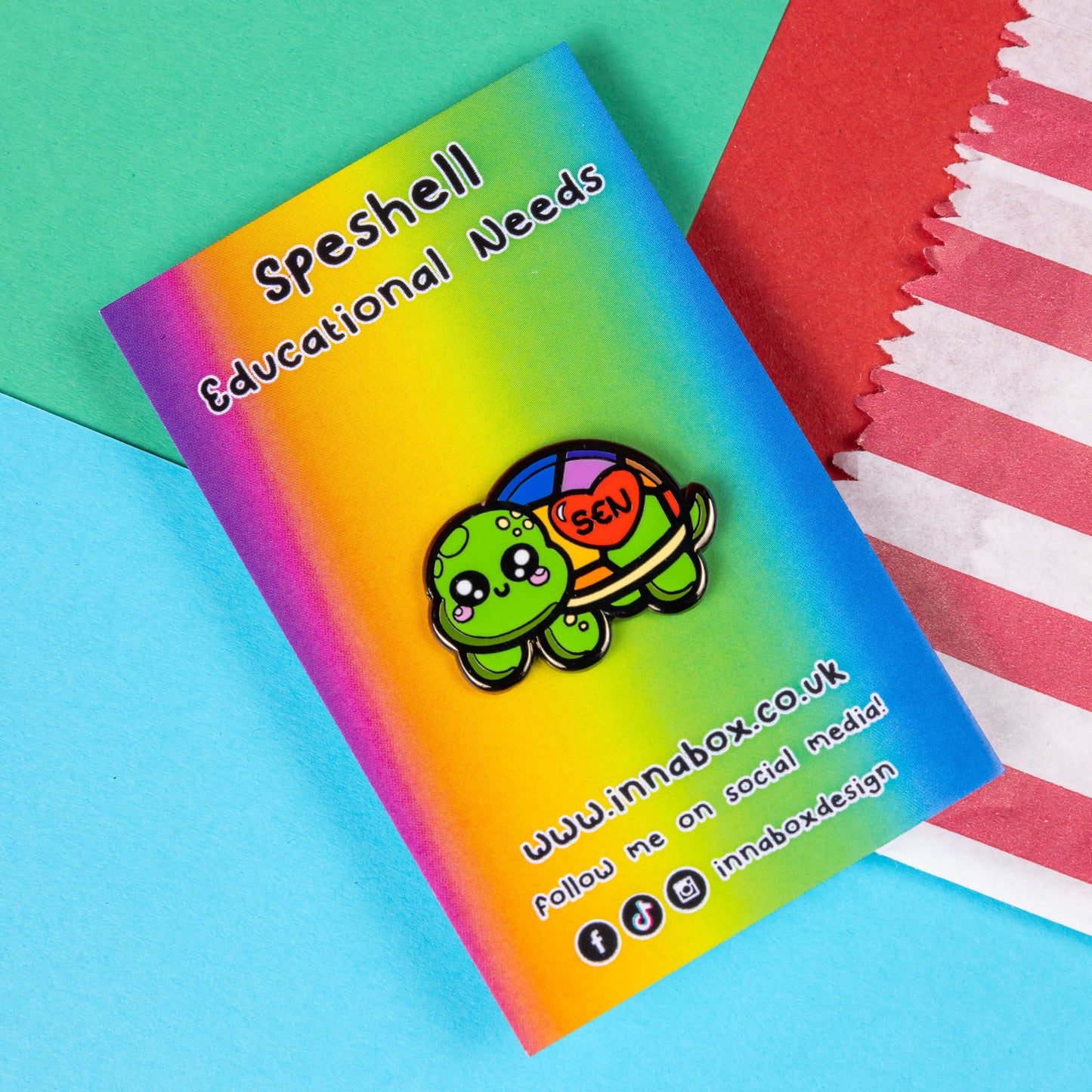 The Speshell Educational Needs Enamel Pin - SEN - Special Educational Needs on a rainbow backing card with black text. A rainbow shell kawaii cute style tortoise with pink cheeks and sparkling eyes, on its shell is a red heart with 'SEN' in the middle. The pin design is raising awareness for SEN Special Educational Needs.