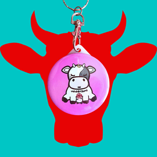 The Uddersons Cow Keyring - Addisons Disease on a red and blue background. The silver clip pink plastic circular keyring features a tired looking black and white cow with a stars above its head, a red swirl on its belly and black text across reading 'uddersons'. The hand drawn design is raising awareness for addisons disease.