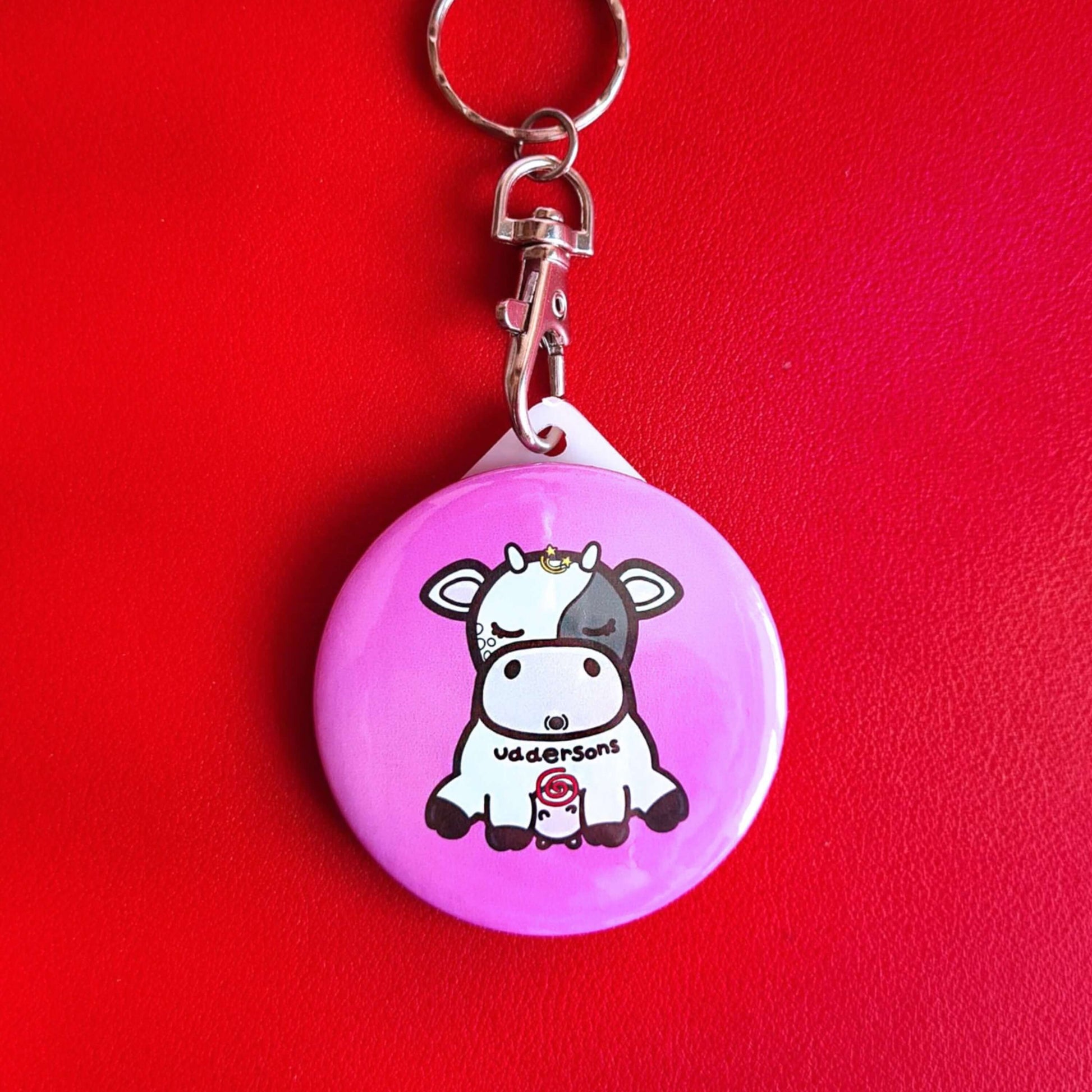 The Uddersons Cow Keyring - Addisons Disease on a red background. The silver clip pink plastic circular keyring features a tired looking black and white cow with a stars above its head, a red swirl on its belly and black text across reading 'uddersons'. The hand drawn design is raising awareness for addisons disease.