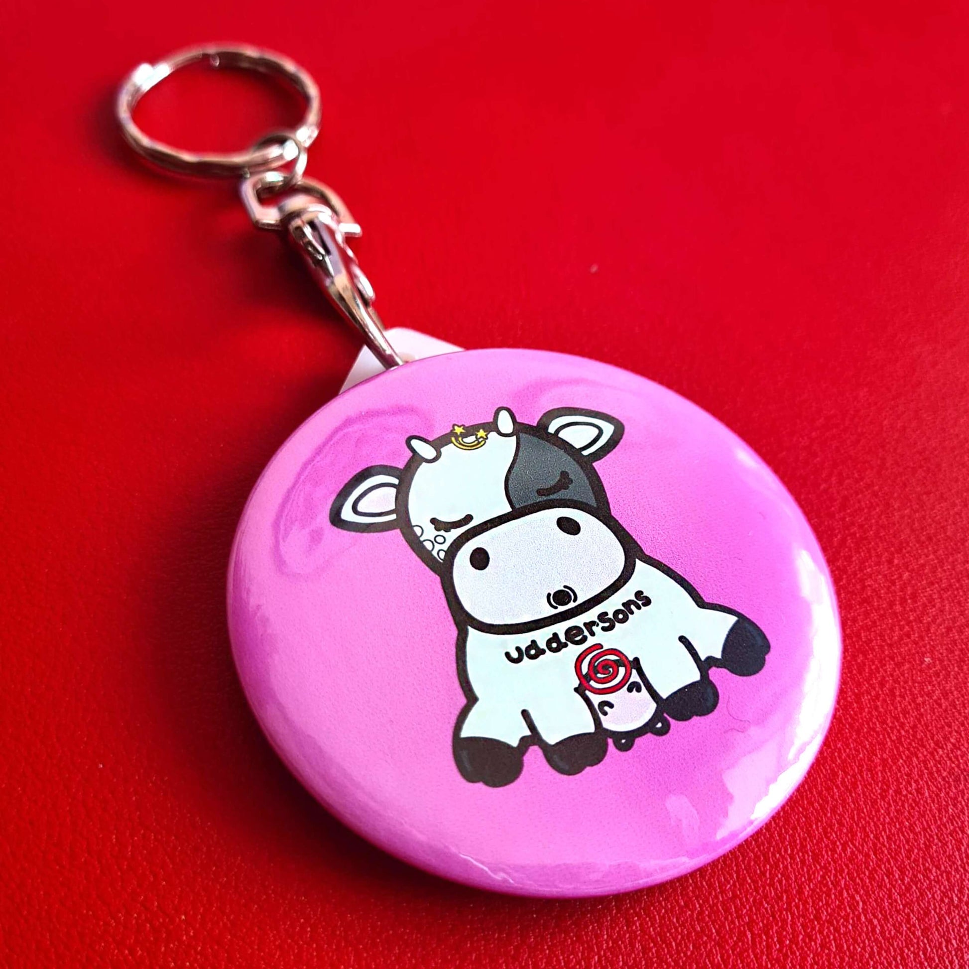 The Uddersons Cow Keyring - Addisons Disease on a red background. The silver clip pink plastic circular keyring features a tired looking black and white cow with a stars above its head, a red swirl on its belly and black text across reading 'uddersons'. The hand drawn design is raising awareness for addisons disease.