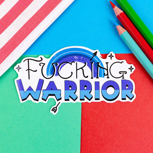 F***ing Warrior Sticker in blue on a red, blue and green background with colouring pencils and red stripe candy bag. The sticker has top swirly black text reading 'fucking' and two tone blue bold bottom text reading 'warrior', behind is a blue rainbow with a black arrow going through and black sparkles surrounding.
