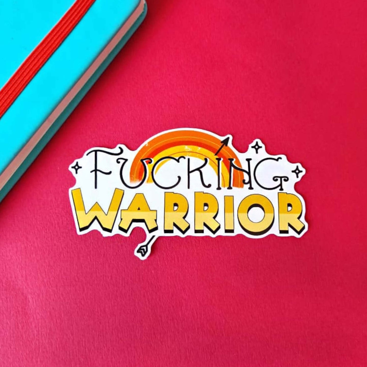 F***ing Warrior Sticker in yellow on a red, blue and green background with colouring pencils and red stripe candy bag. The sticker has top swirly black text reading 'fucking' and two tone yellow bold bottom text reading 'warrior', behind is a yellow rainbow with a black arrow going through and black sparkles surrounding.