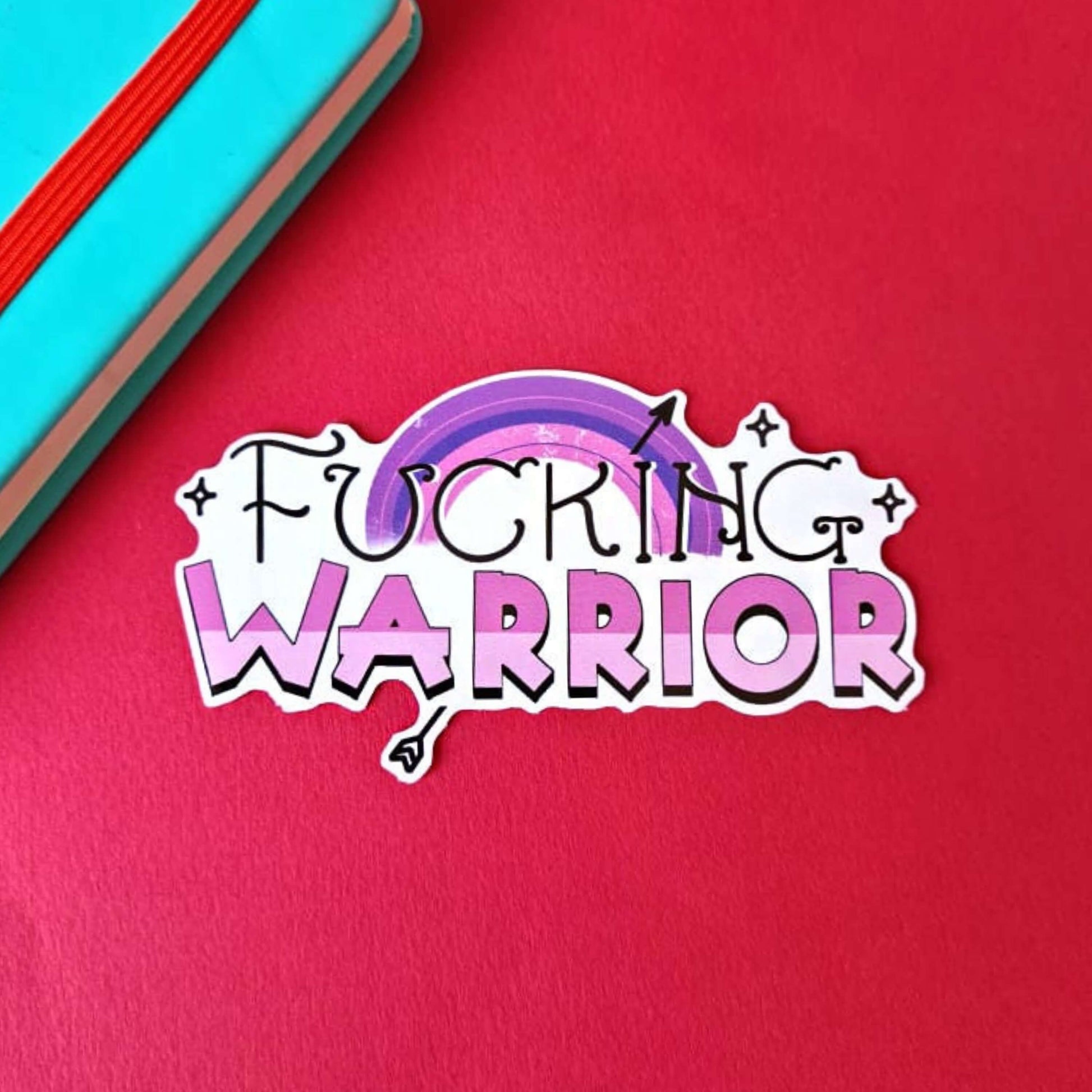 F***ing Warrior Sticker in purple on a red, blue and green background with colouring pencils and red stripe candy bag. The sticker has top swirly black text reading 'fucking' and two tone purple bold bottom text reading 'warrior', behind is a purple rainbow with a black arrow going through and black sparkles surrounding.