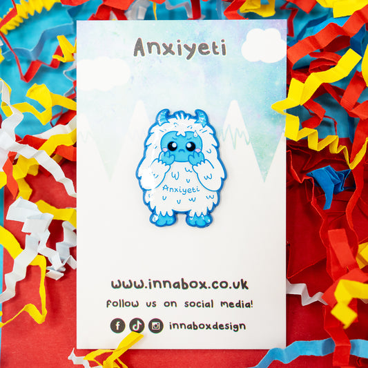 Anxi-Yeti hard enamel pin by Innabox, showing a cute blue yeti character with glossy detailing and the word “Anxiyeti” on its chest. Displayed on an illustrated mountain-themed backing card with bright confetti. A kind, disability-inspired pin designed to comfort and raise awareness.
