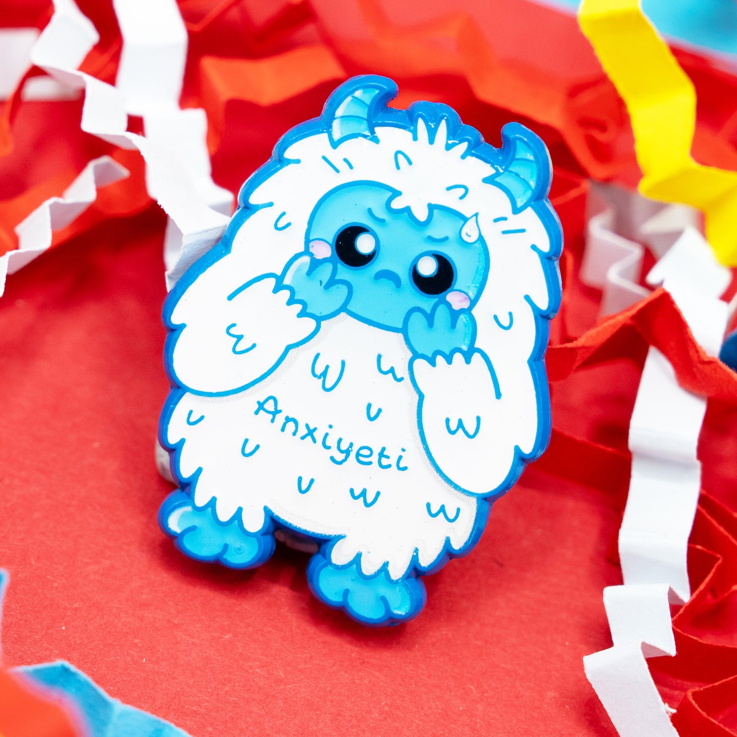Close-up of the Anxi-Yeti enamel pin by Innabox, a sweet blue and white creature with a nervous yet adorable face. Surrounded by colourful shredded paper on a red background. A mental-health awareness pin designed to uplift those with anxiety, chronic illness and invisible disabilities.