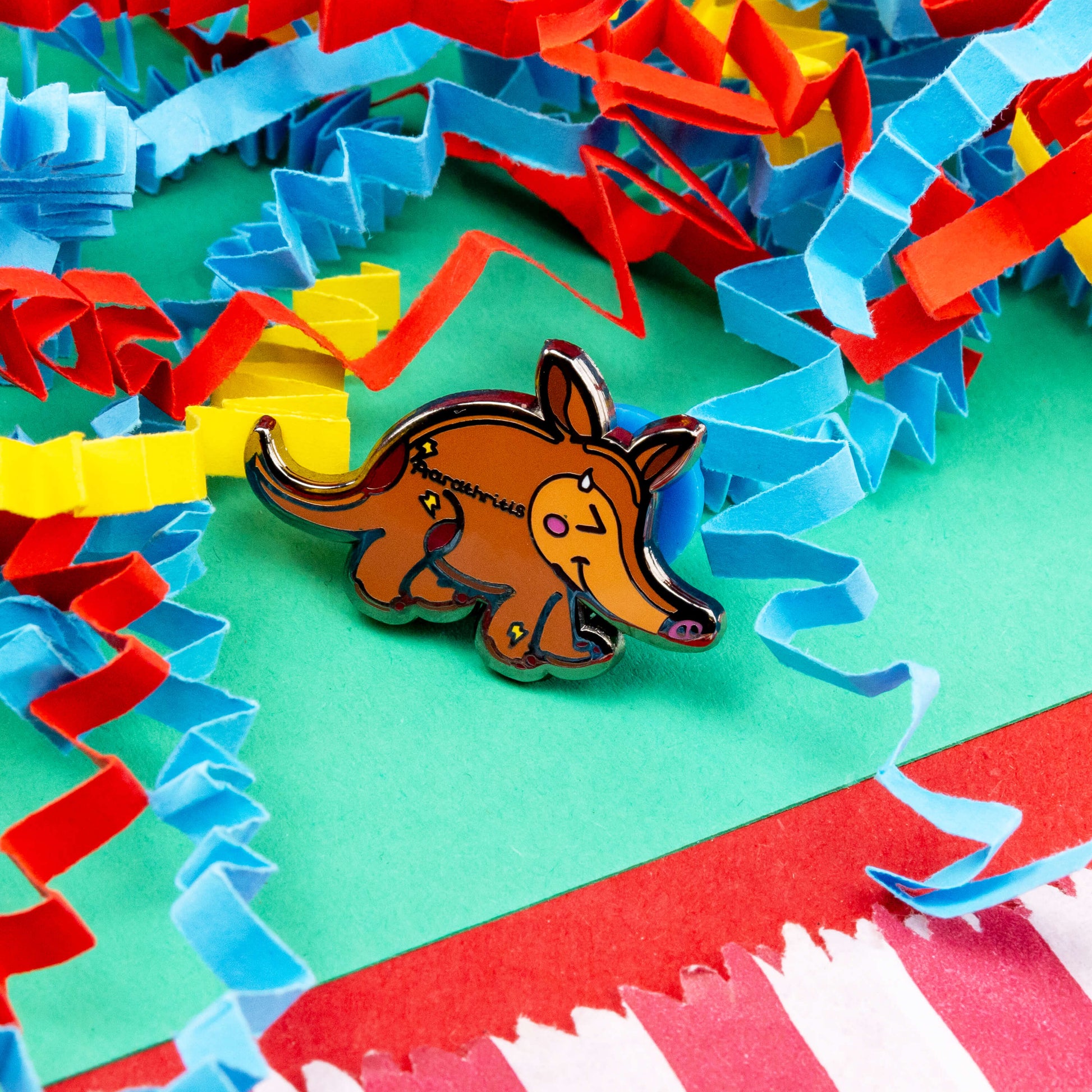 The Aardthritis Aardvark Enamel Pin - Arthritis on a red, blue, green and yellow crinkle card confetti background. The smiling sweating brown aardvark with pink cheeks and lightning bolts scattered across its body with black text reading 'aardthritis'. The hand drawn design is raising awareness for arthritis, Osteoarthritis and Rheumatoid arthritis.