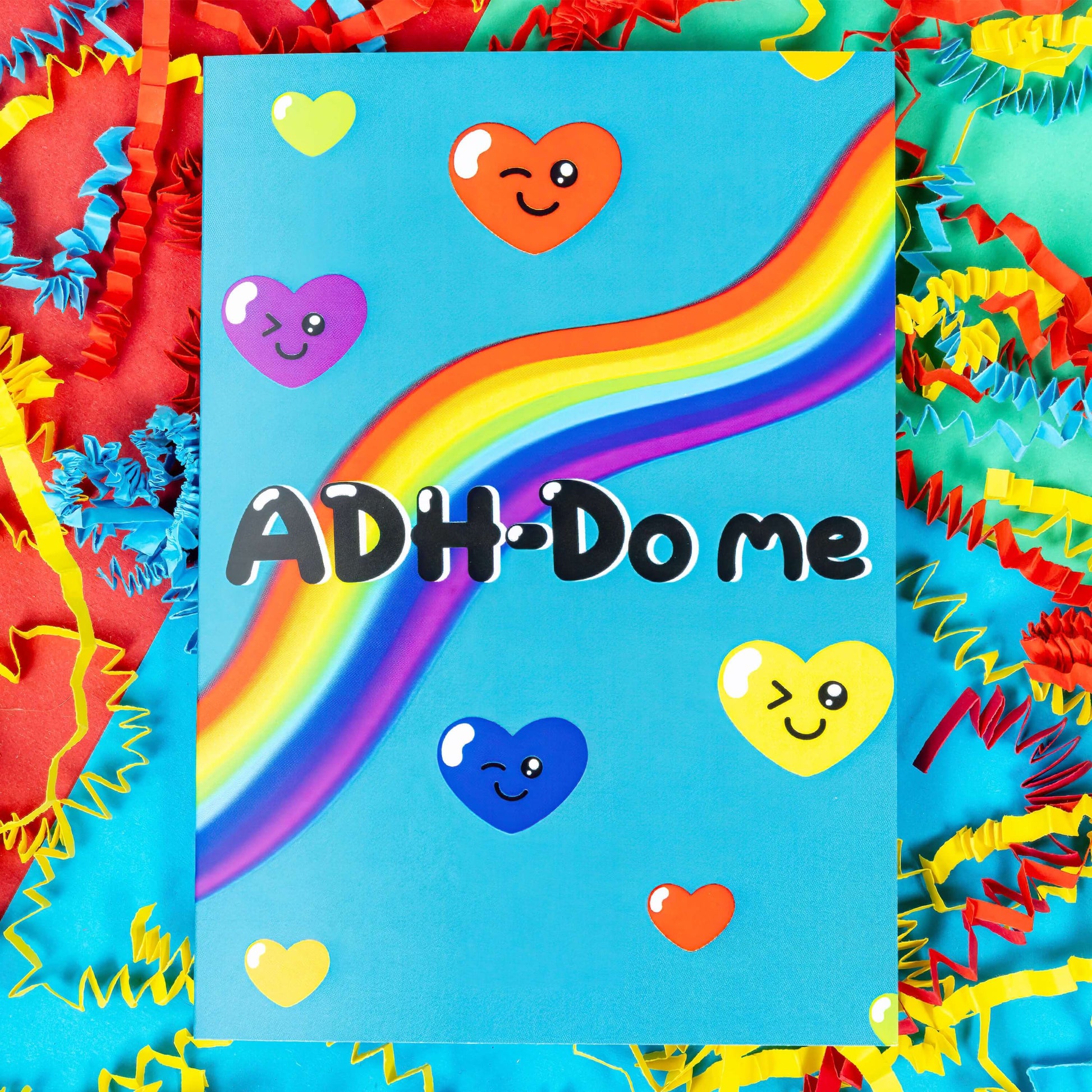 A sky blue greeting card with a rainbow going across it with different coloured love hearts with winky faces dotted around it. 'ADH-Do me' is written in big black letters across the card. The background of the photo is multi coloured card confetti. Design is inspired by neurodivergent disorders.