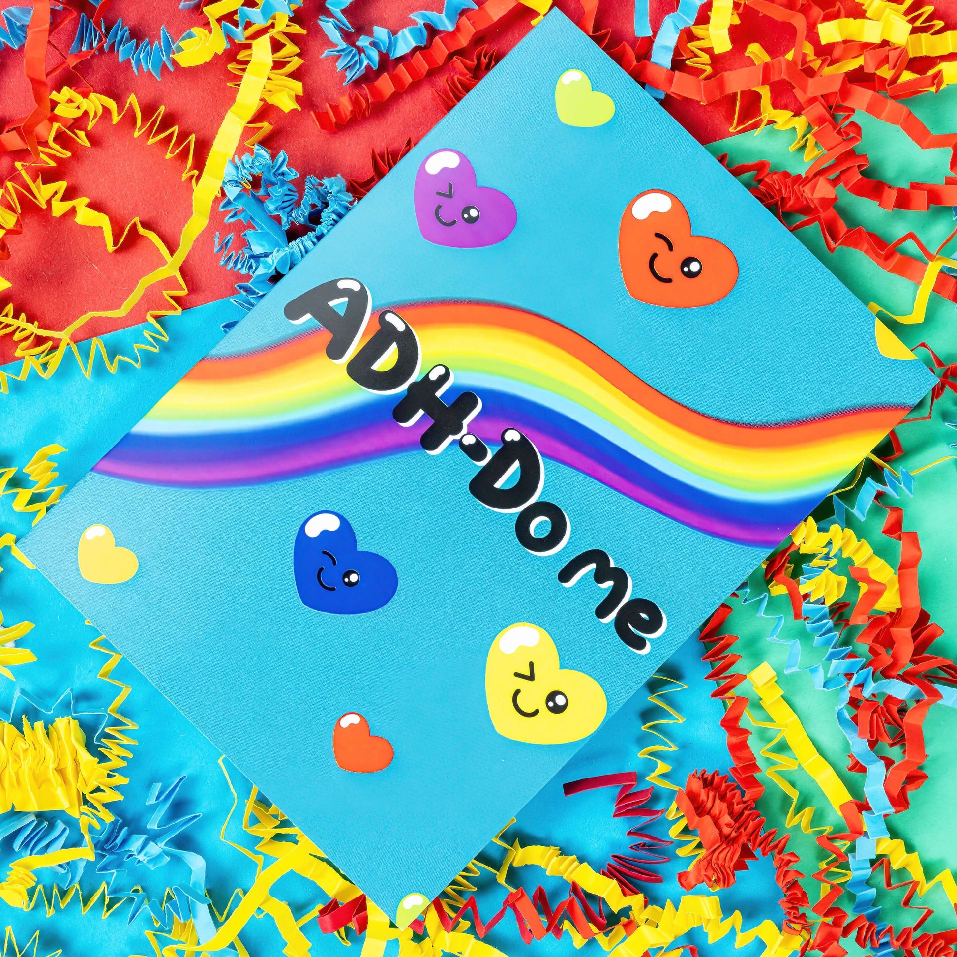 A sky blue greeting card with a rainbow going across it with different coloured love hearts with winky faces dotted around it. 'ADH-Do me' is written in big black letters across the card. The background of the photo is multi coloured card confetti. Inspired by neurodivergent disorders.