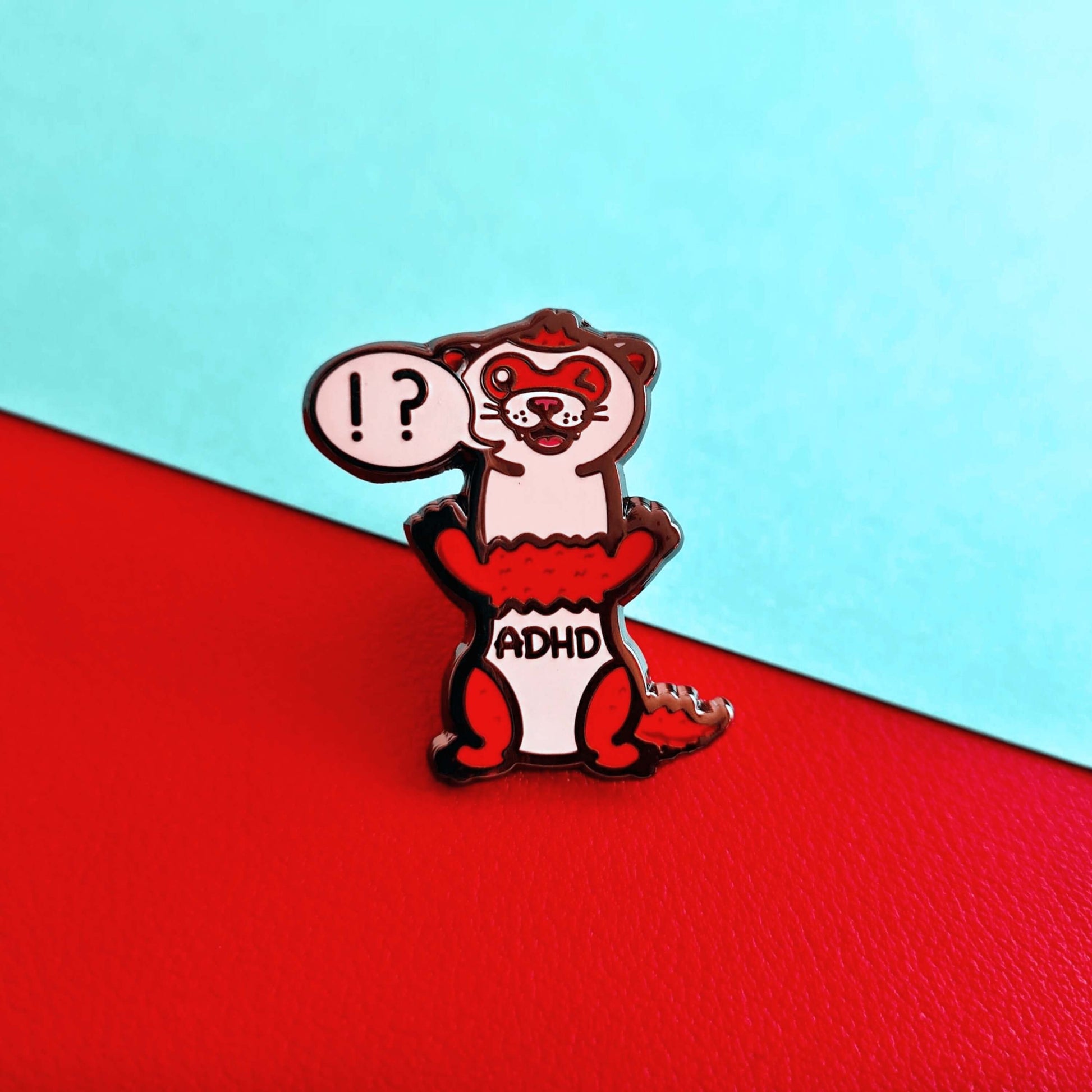 A red and white ferret enamel pin with it's mouth open in a smile and it's little teeth are showing. One of it's eyes are closed shut and it is standing on it's back legs. There is a speech-bubble from the ferret's mouth with '!?' inside it. The enamel pin is shown on a red and blue background. Raising awareness for ADHD - Attention deficit hyperactivity disorder.