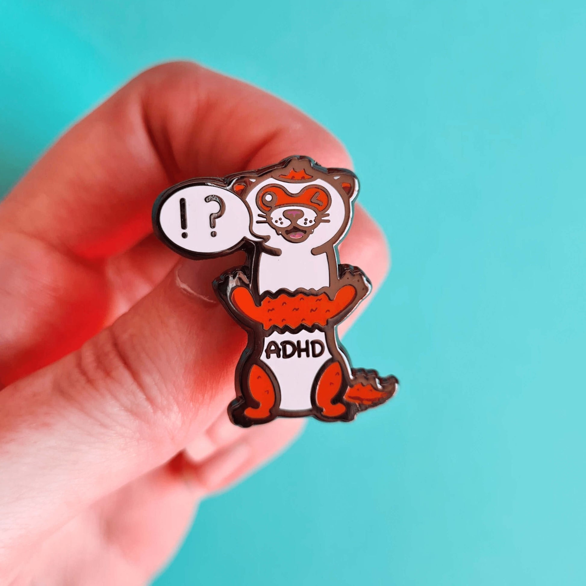 A red and white ferret enamel pin with it's mouth open in a smile and it's little teeth are showing. One of it's eyes are closed shut and it is standing on it's back legs. There is a speech-bubble from the ferret's mouth with '!?' inside it. The enamel pin is being held over a blue background. Raising awareness for ADHD - Attention deficit hyperactivity disorder.