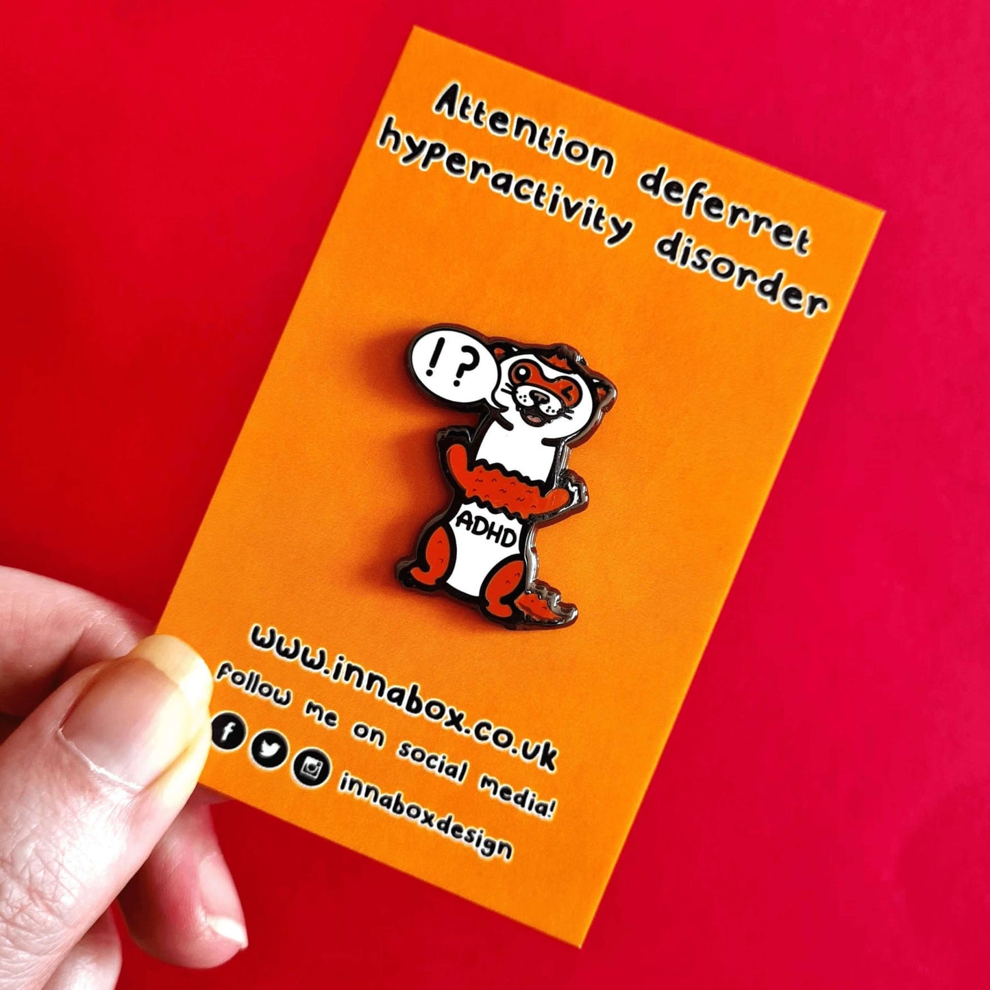 A red and white ferret enamel pin with it's mouth open in a smile and it's little teeth are showing. One of it's eyes are closed shut and it is standing on it's back legs. There is a speech-bubble from the ferret's mouth with '!?' inside it. The enamel pin is being held over a red background on orange backing card. Raising awareness for ADHD - Attention deficit hyperactivity disorder.