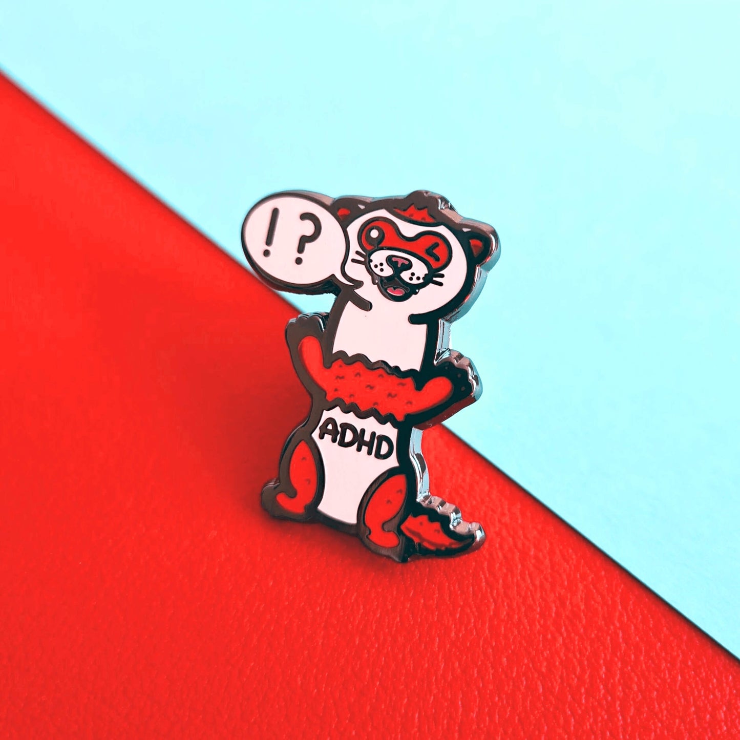 A red and white ferret enamel pin with it's mouth open in a smile and it's little teeth are showing. One of it's eyes are closed shut and it is standing on it's back legs. There is a speech-bubble from the ferret's mouth with '!?' inside it. The enamel pin is shown on a red and blue background. Raising awareness for ADHD - Attention deficit hyperactivity disorder.