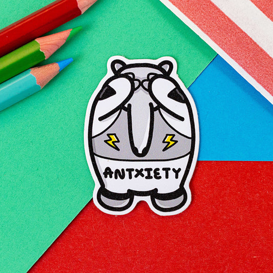 Antxiety Sticker - Anxiety on a red, blue and green background with a red stripe candy bag and colouring pencils. The sticker is a grey and white anteater with yellow lightning bolts and the word antxiety across its belly. The sticker is designed to raise awareness for anxiety disorders and anxious emotions.