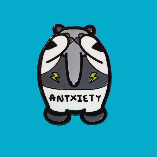 Antxiety Enamel Pin - Anxiety sat on a blue background. The pin is a grey and white anteater with yellow lightning bolts and the word antxiety across its belly. The pin is designed to raise awareness for anxiety disorders and anxious emotions.