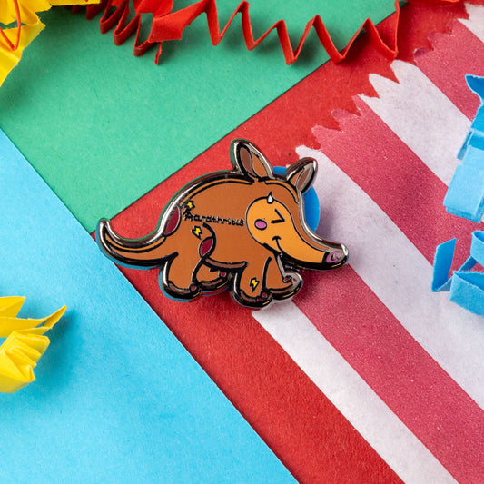 The Aardthritis Aardvark Enamel Pin - Arthritis on a red, blue, green and yellow crinkle card confetti background. The smiling sweating brown aardvark with pink cheeks and lightning bolts scattered across its body with black text reading 'aardthritis'. The hand drawn design is raising awareness for arthritis, Osteoarthritis and Rheumatoid arthritis.