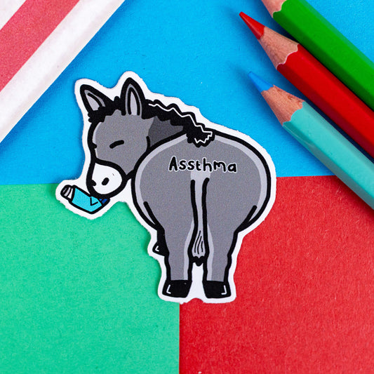 Assthma Sticker - Asthma on a blue, red and green background with colouring pencils and a red stripe candy bag. A grey donkey ass showing its behind with a blue asthma pump in its mouth and the word Assthma across its behind. The sticker is designed to raise awareness for asthma.