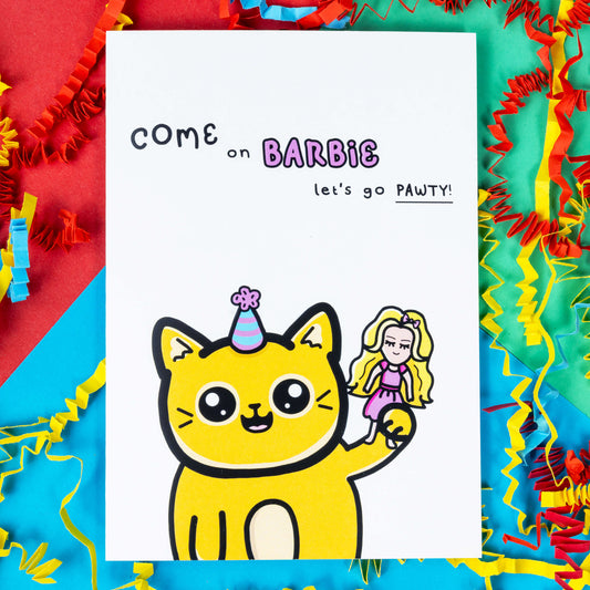 The Let's Go Pawty Cat Birthday Card on a blue, red and green background with yellow, blue and red crinkle card confetti. The white background a6 card has a yellow smiling cat with a pastel party hat holding up a pink dressed doll with blonde hair. Above the cat in black text reads 'come on barbie let's go pawty!'. The barbie section of the text is pink bubble writing.