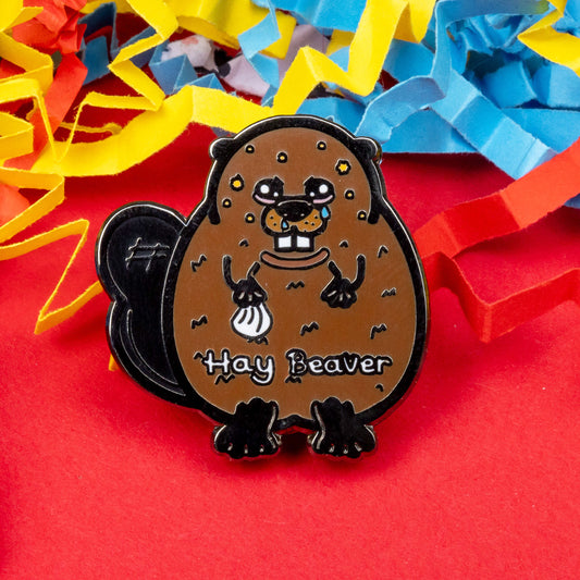 Hay Beaver Enamel Pin - Hay Fever on a blue, yellow and red coloured card confetti background. The enamel pin is a brown beaver with watery eyes, dripping nose and yellow spots on face and is holding a tissue with it's little hand. Hay beaver is written across its belly. The enamel pin is designed to raise awareness for hay fever or allergic rhinitis