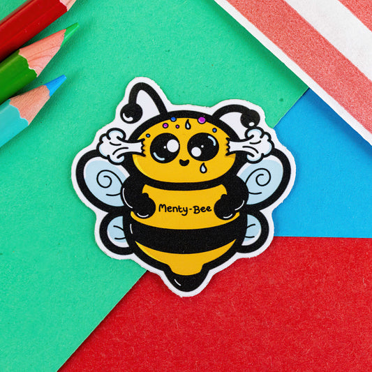 The Menty Bee Sticker - Mental Breakdown on a red, blue and green background with colouring pencils and red stripe candy bag. The stressed bumble bee shape sticker has steaming ears, lopsided eyes, sweat drops and multicoloured spots on its head. Across its middle in black text reads 'menty-bee'. The hand drawn design is raising awareness for mental breakdowns and mental health.