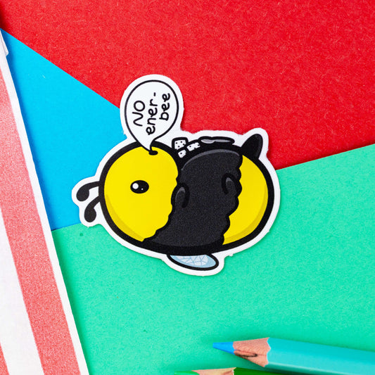 The Low Energy Bee Sticker on a red, blue and green background with colouring pencils and red stripe candy bag. The bumble bee shaped vinyl sticker is laying on its back with sugar cubes and a spoon on its belly with a speech bubble reading 'no ener-bee'. The hand drawn design is raising awareness for chronic fatigue.