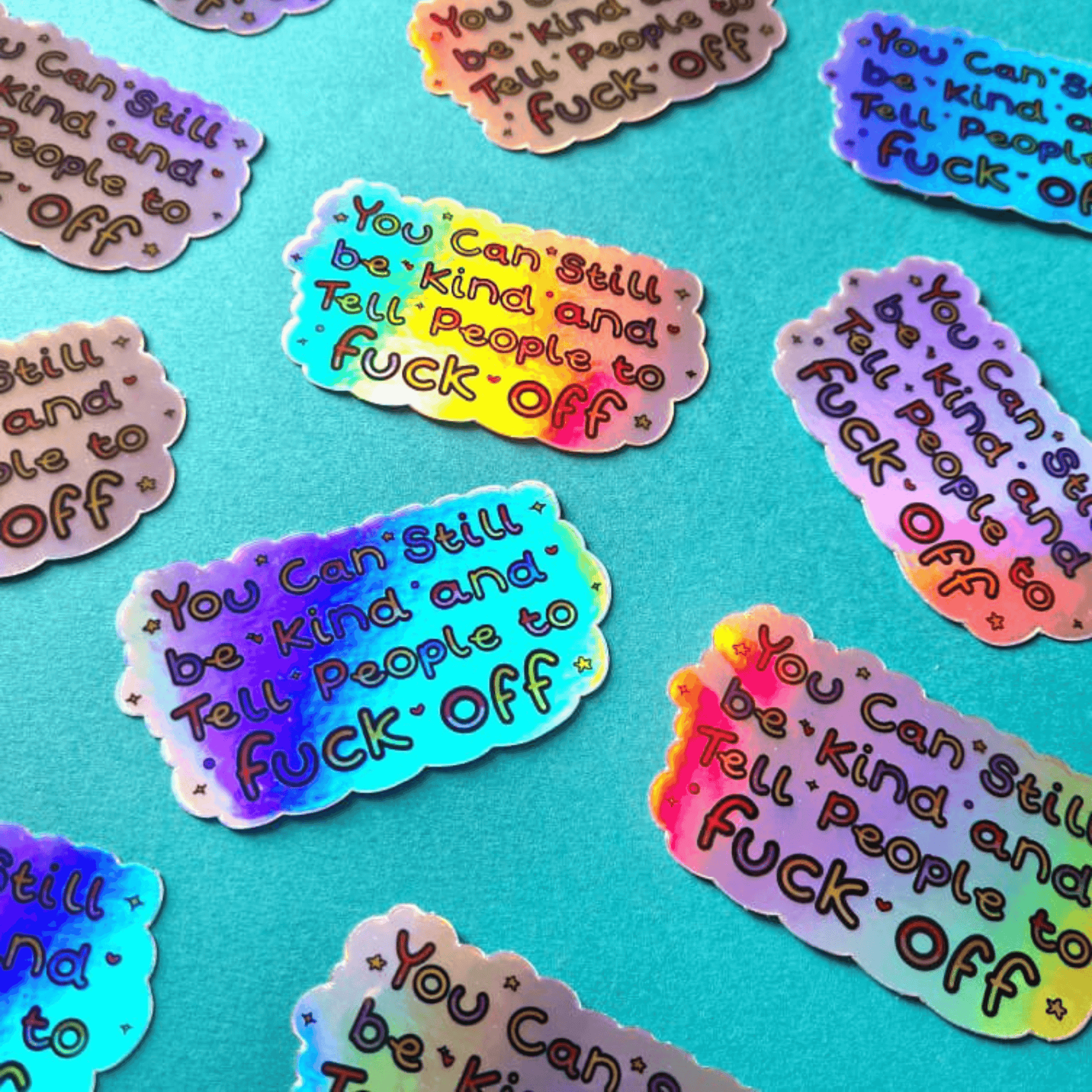 Multiple You Can Still be Kind and Tell People to F**k Off Stickers on a blue background. The sticker is holographic with rainbow writing with sparkles, hearts, stars and circles.