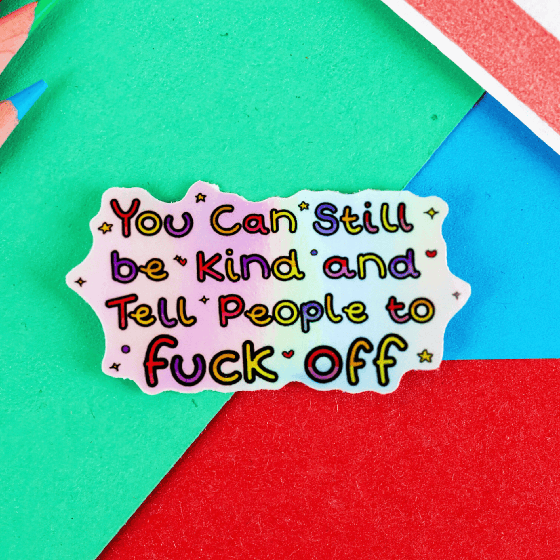 You Can Still be Kind and Tell People to F**k Off Sticker on a green, blue and red background with colouring pencils and a red stripe candy bag. The sticker is holographic with rainbow writing with sparkles, hearts, stars and circles. 