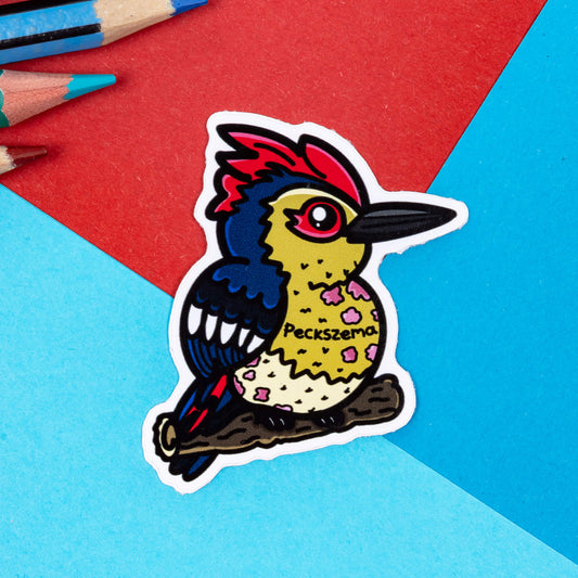 The Peckszema Woodpecker Sticker - Eczema on a red and blue background with colouring pencils. The blue, red and yellow woodpecker bird has pink dry patches all over its body with black text reading 'peckszema'. The hand drawn design is raising awareness for eczema.