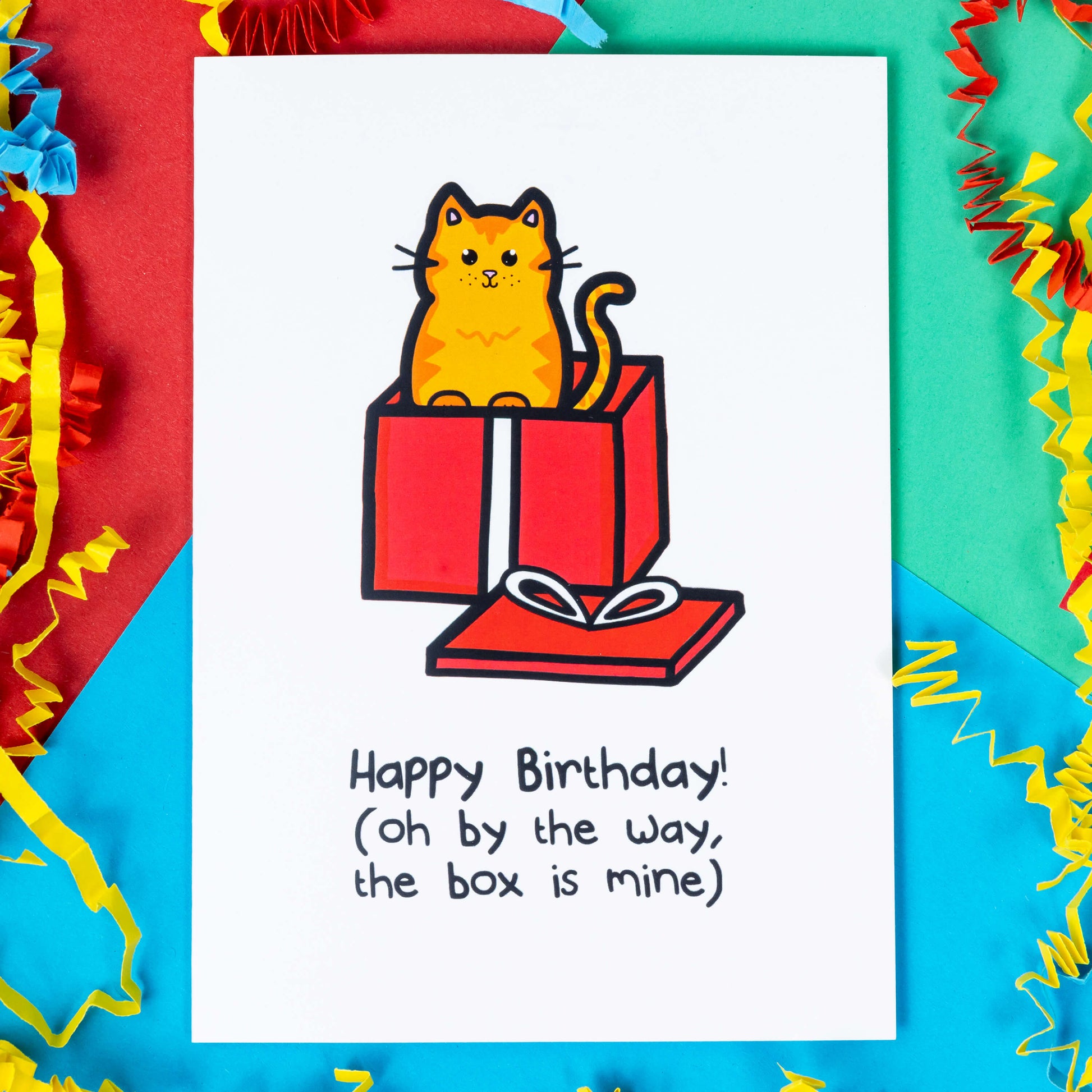 A white card with a ginger cat illustration coming out of a red present with text saying Happy Birthday! (Oh by the way, the box is mine). The a6 birthday card is laying on a red, blue and green background with red, yellow and blue crinkle card confetti.