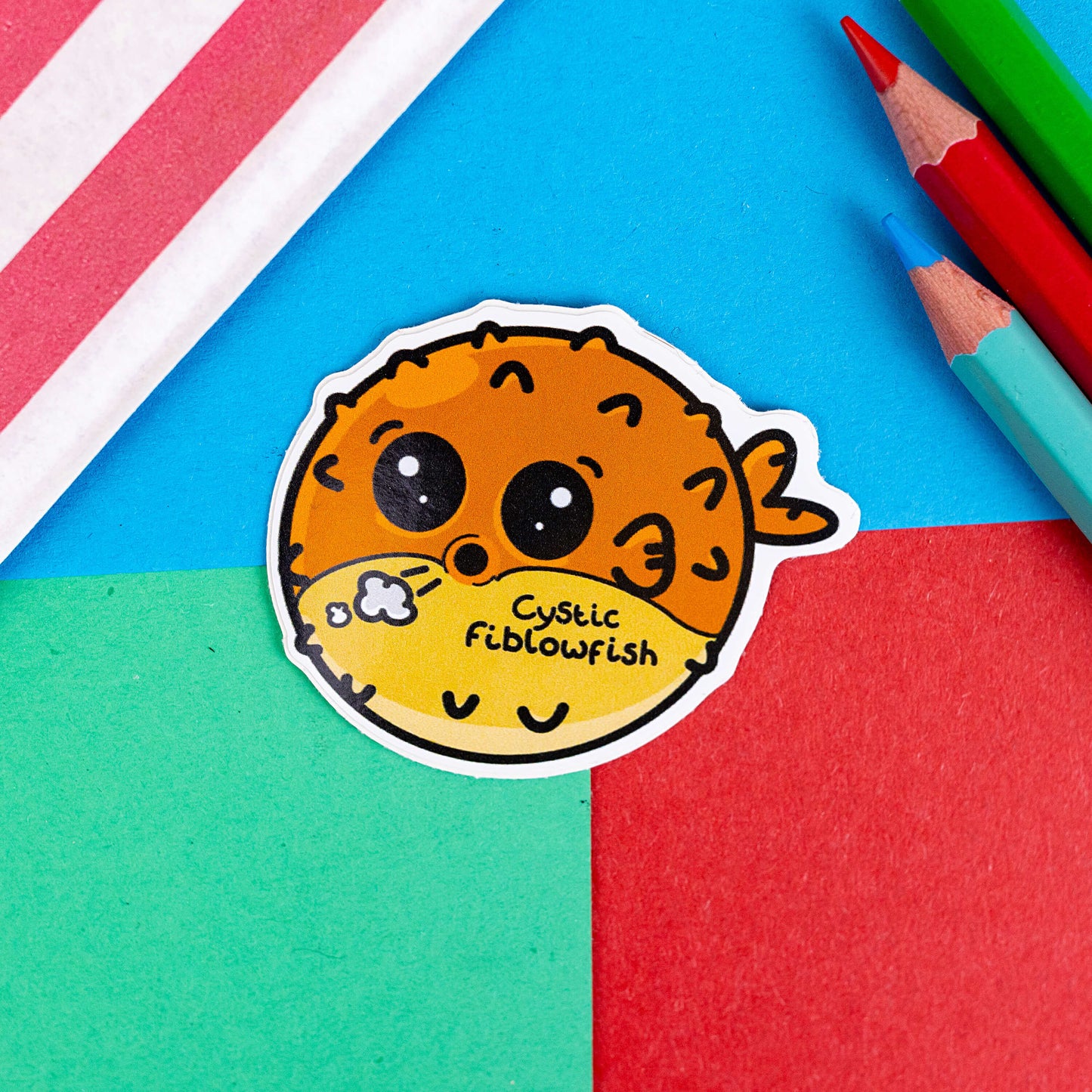 The Cystic Fiblowfish Sticker - Cystic Fibrosis on a red, blue and green background with colouring pencils and red stripe candy bag. The pufferfish shape sticker is wheezing with clouds coming out its mouth and black text across its belly reading 'cystic fiblowfish'. The design is raising awareness for cystic fibrosis.
