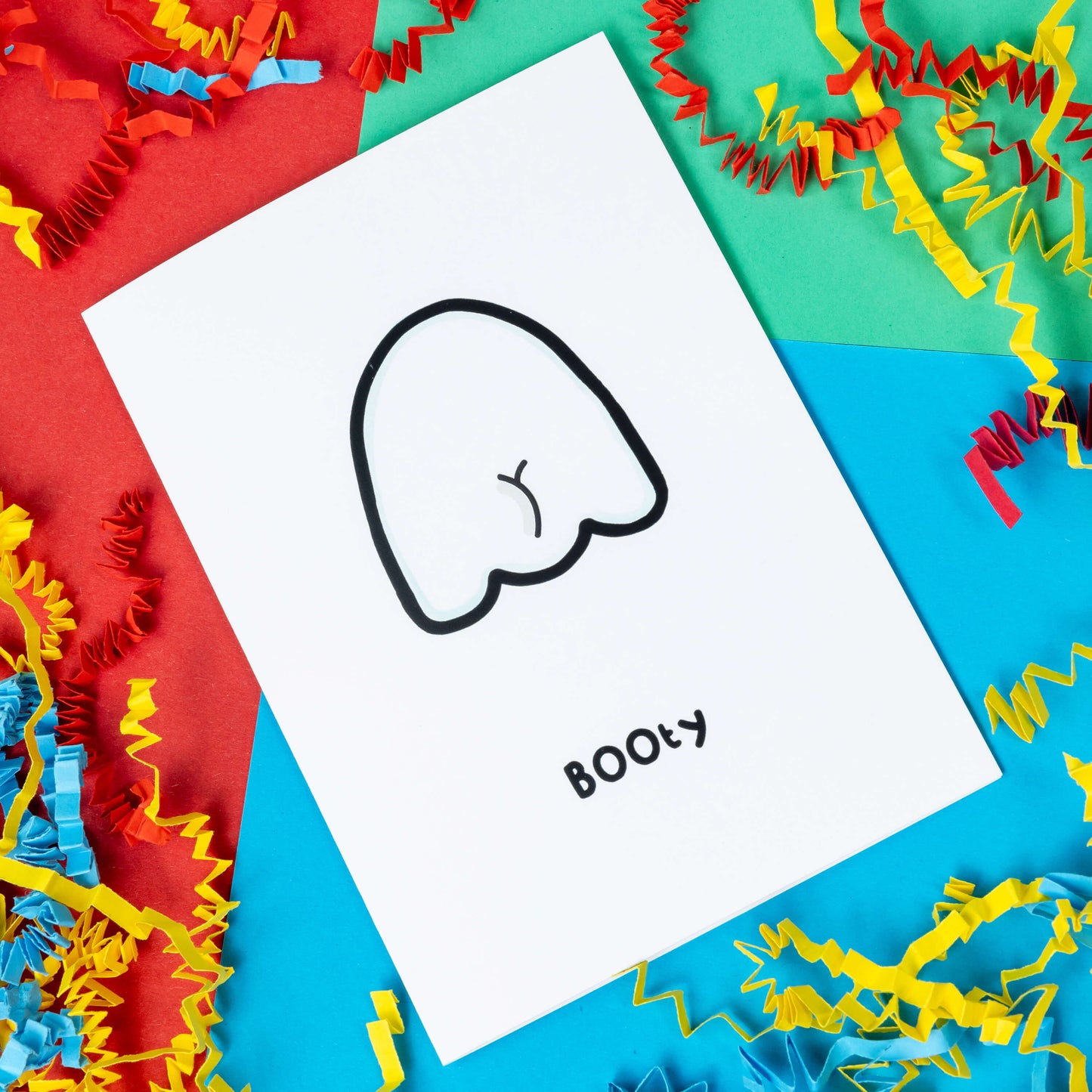The Booty Ghost a6 card on a blue, red and green background with yellow, blue and red crinkle card confetti. A white greeting card with an illustration of the back of a ghost with a bum crack. The word 'BOOty" is written in black underneath the drawing.