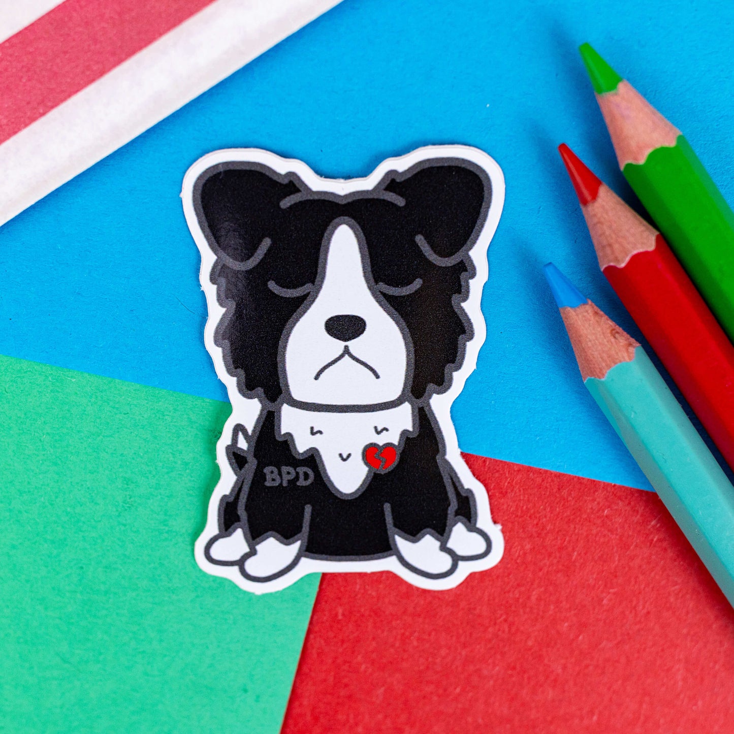 BPD Sticker - Borderline Personality Disorder on a blue, red and green background with colouring pencils and a red stripe candy bag. The black and white sad fluffy dog sticker has a broken heart with BPD written across its chest. The sticker is designed to raise awareness for Borderline Personality Disorder.