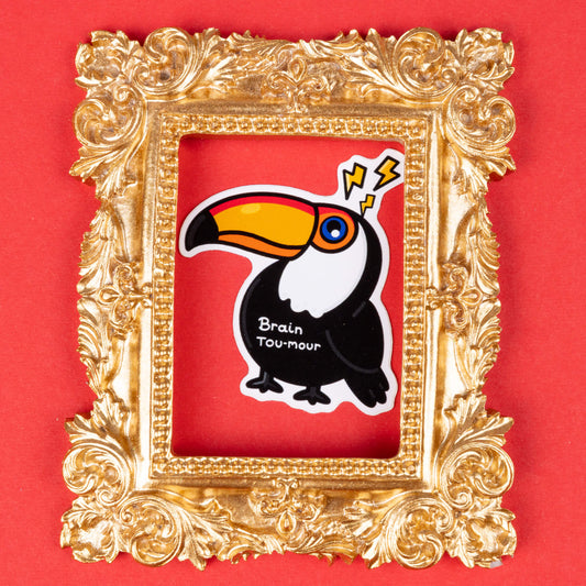 A sticker of a toucan with a bright orange beak and black and white feathers, with lightning bolts above its head. The words "Brain Tou-mour" are written on its chest. The sticker is framed in an ornate gold picture frame and set against a red background. Hand drawn design created to raise awareness for brain tumours.