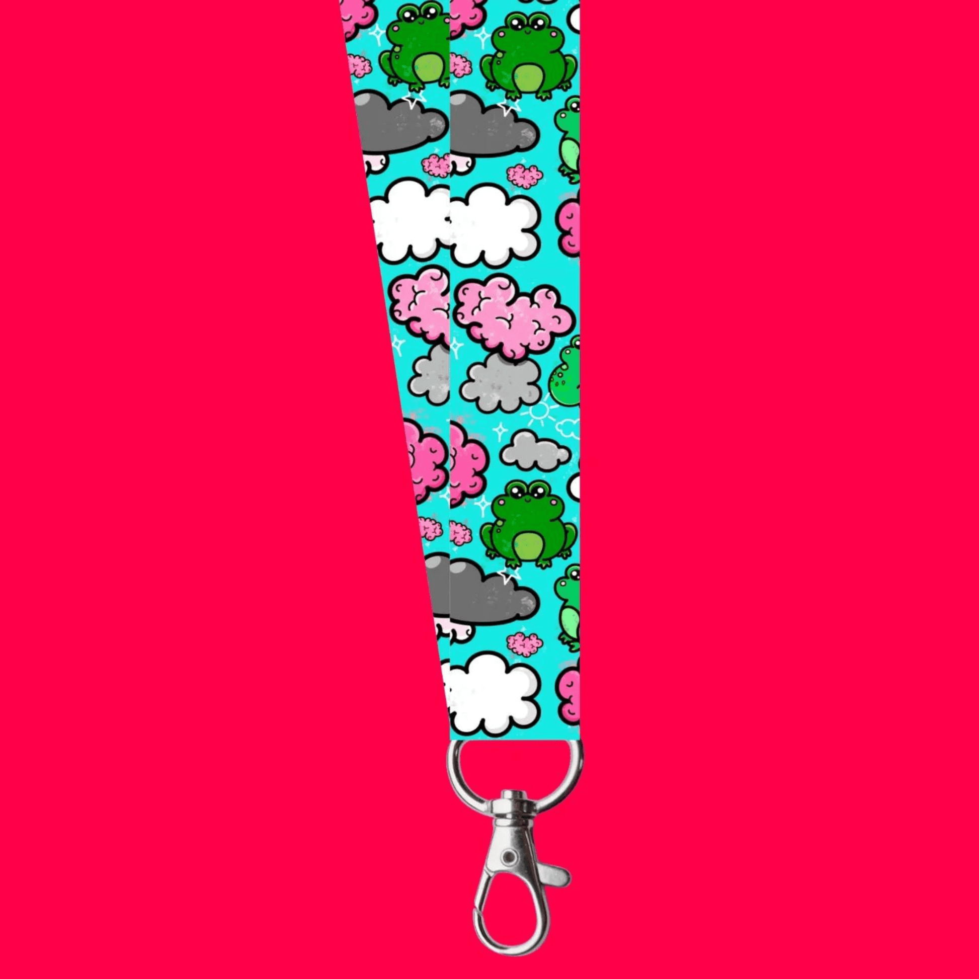 The Brain Frog Lanyard - Brain Fog on a red background. The lanyard has a silver lobster clip with a blue base covered in various kawaii face green frogs with pink cheeks, pink brain styled clouds with white and grey clouds and white sparkles all over. The design was created to raise awareness for brain fog.