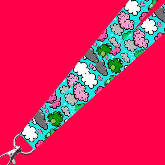 The Brain Frog Lanyard - Brain Fog on a red background. The lanyard has a silver lobster clip with a blue base covered in various kawaii face green frogs with pink cheeks, pink brain styled clouds with white and grey clouds and white sparkles all over. The design was created to raise awareness for brain fog.
