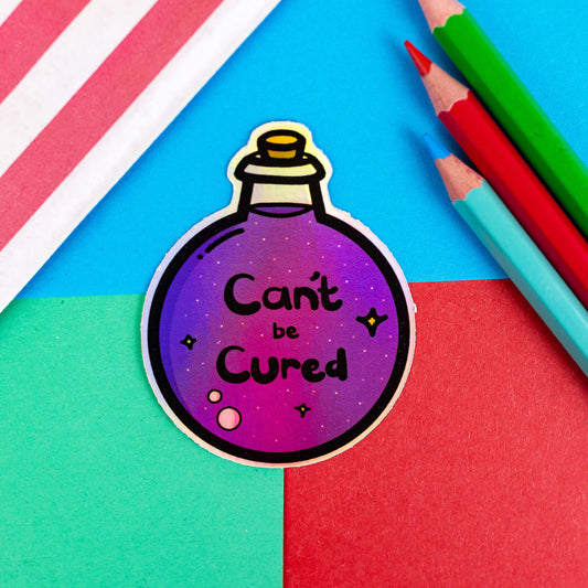 The Can't Be Cured Holographic Sticker on a red, blue and green background with colouring pencils and red stripe candy bag. The sticker is a circular potion bottle with a cork stop top and purple middle. In the middle is white and yellow sparkles, two pastel pink bubbles and black text that reads 'can't be cured'. The design was inspired by chronic illnesses.