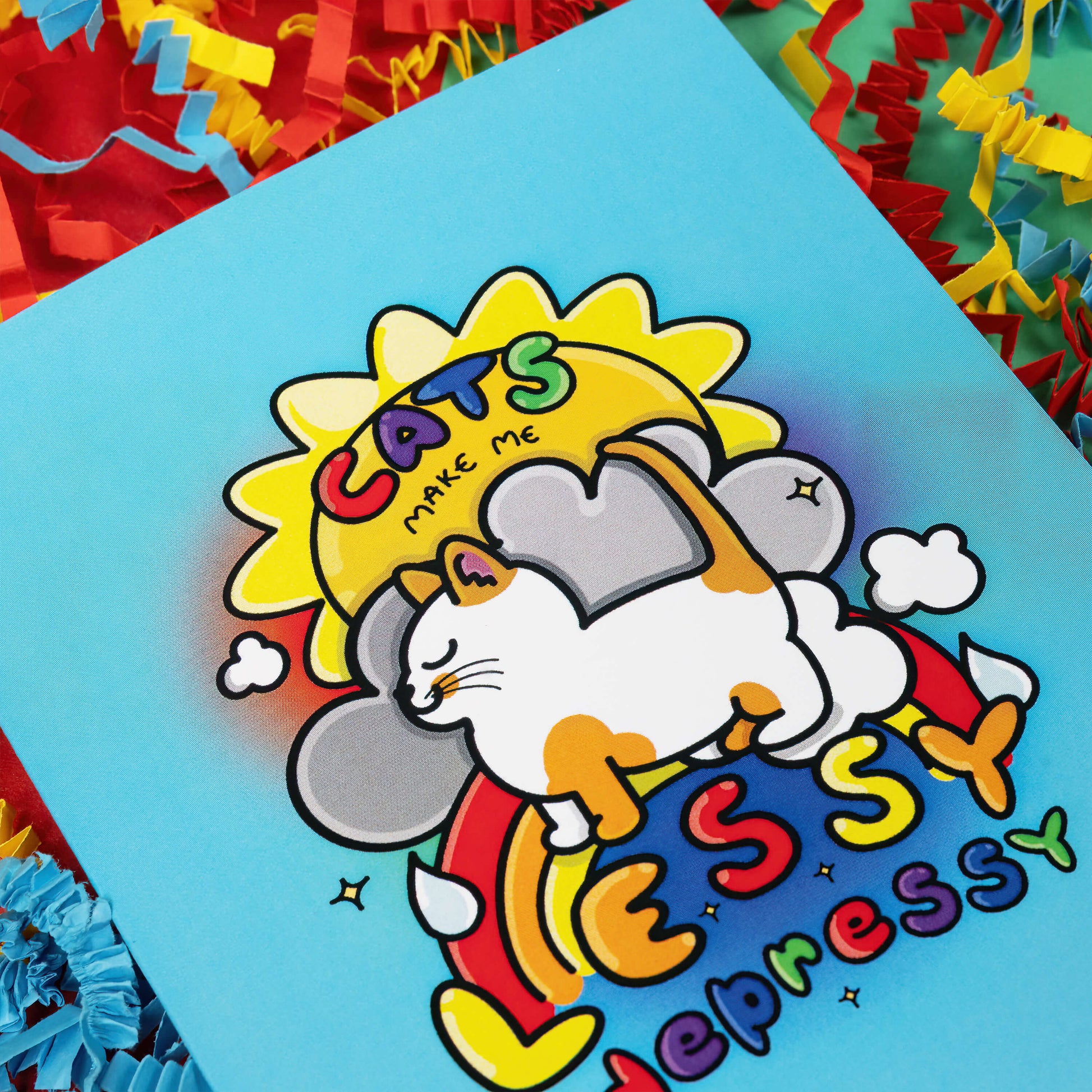 Close up of a light blue greeting card with an illustration of a happy ginger and white cat with a sun, clouds and rainbow behind it. 'Cats make me lessy depressy' is written on the card in rainbow bubble writing. The background of the photo is red, blue and yellow crinkle card confetti.