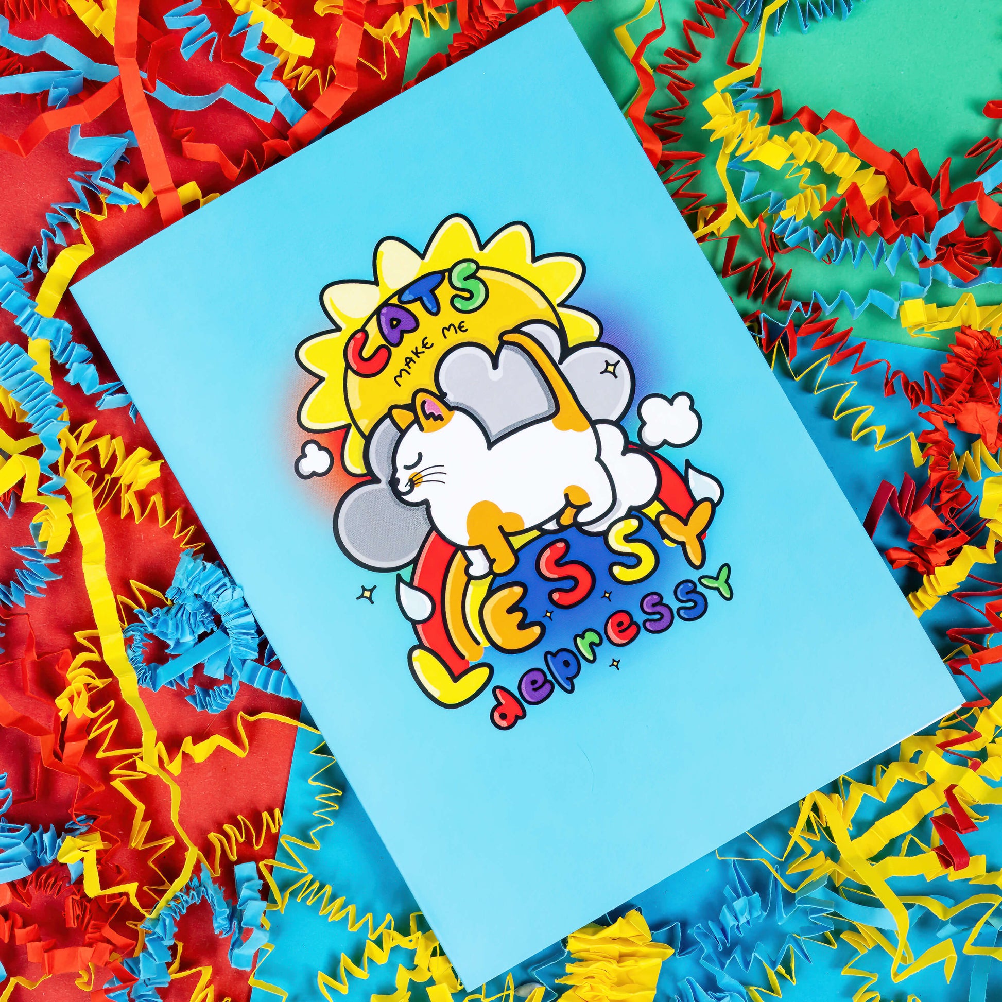 A light blue greeting card with an illustration of a happy ginger and white cat with a sun, clouds and rainbow behind it. 'Cats make me lessy depressy' is written on the card in rainbow bubble writing. The background of the photo is red, blue and yellow crinkle card confetti.
