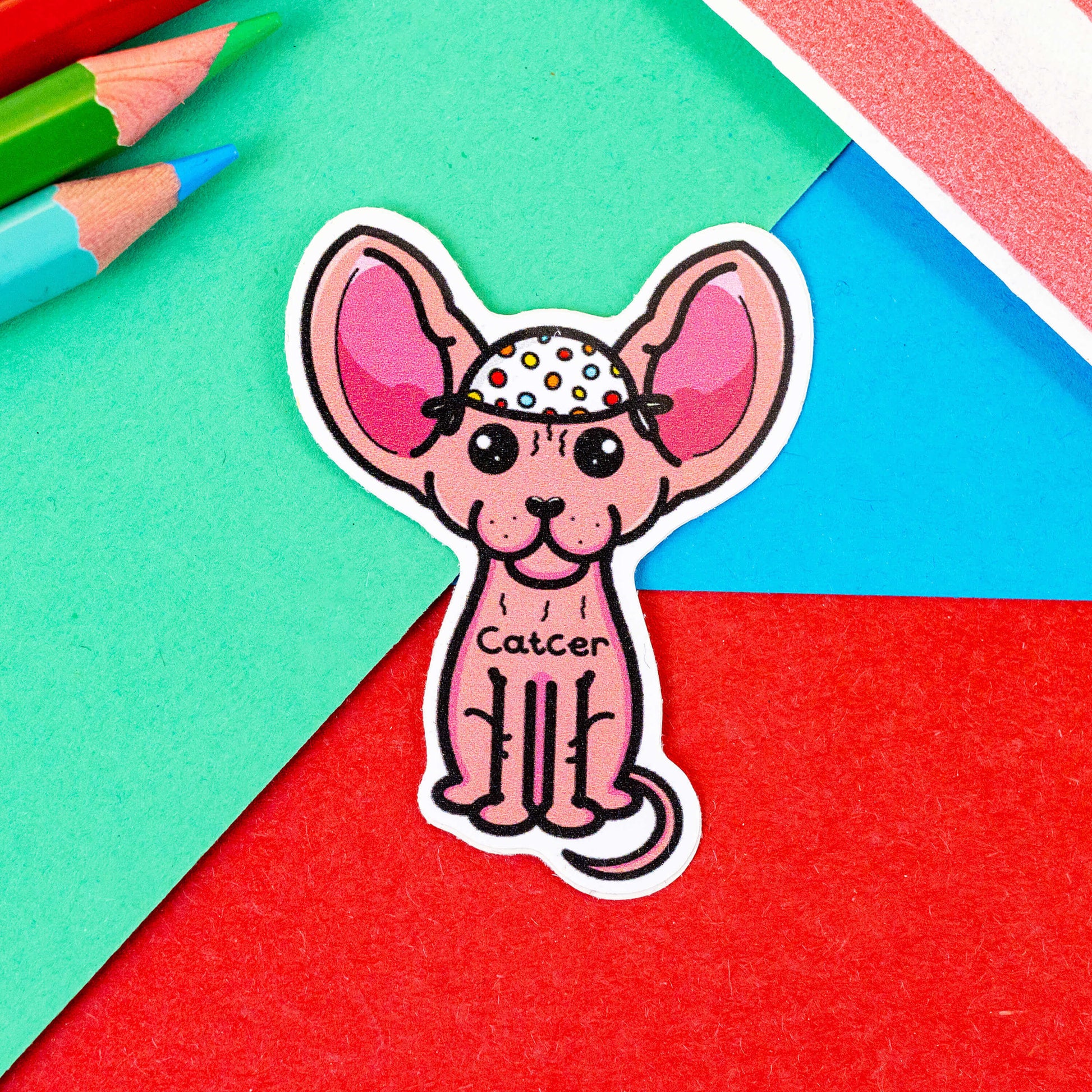The Catcer Cat Sticker - Cancer on a red, green and blue background with colouring pencils and a red stripe candy bag. The sticker is a pink hairless sphynx cat wearing a white medical cap with rainbow polka dots, across its chest reads 'Catcer'. Designed to raise awareness for cancer.