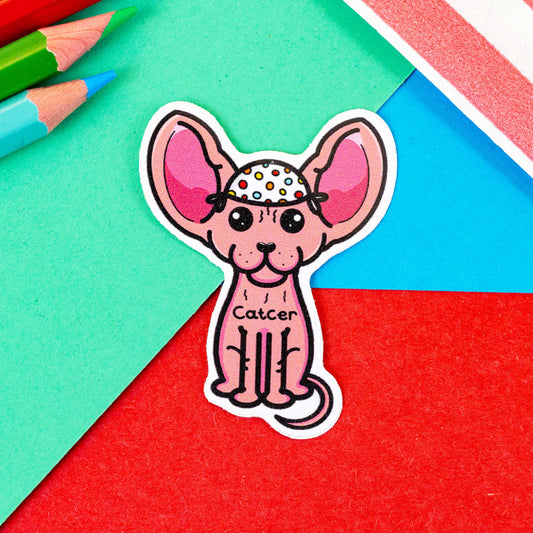 The Catcer Cat Sticker - Cancer on a red, green and blue background with colouring pencils and a red stripe candy bag. The sticker is a pink hairless sphynx cat wearing a white medical cap with rainbow polka dots, across its chest reads 'Catcer'. Designed to raise awareness for cancer.