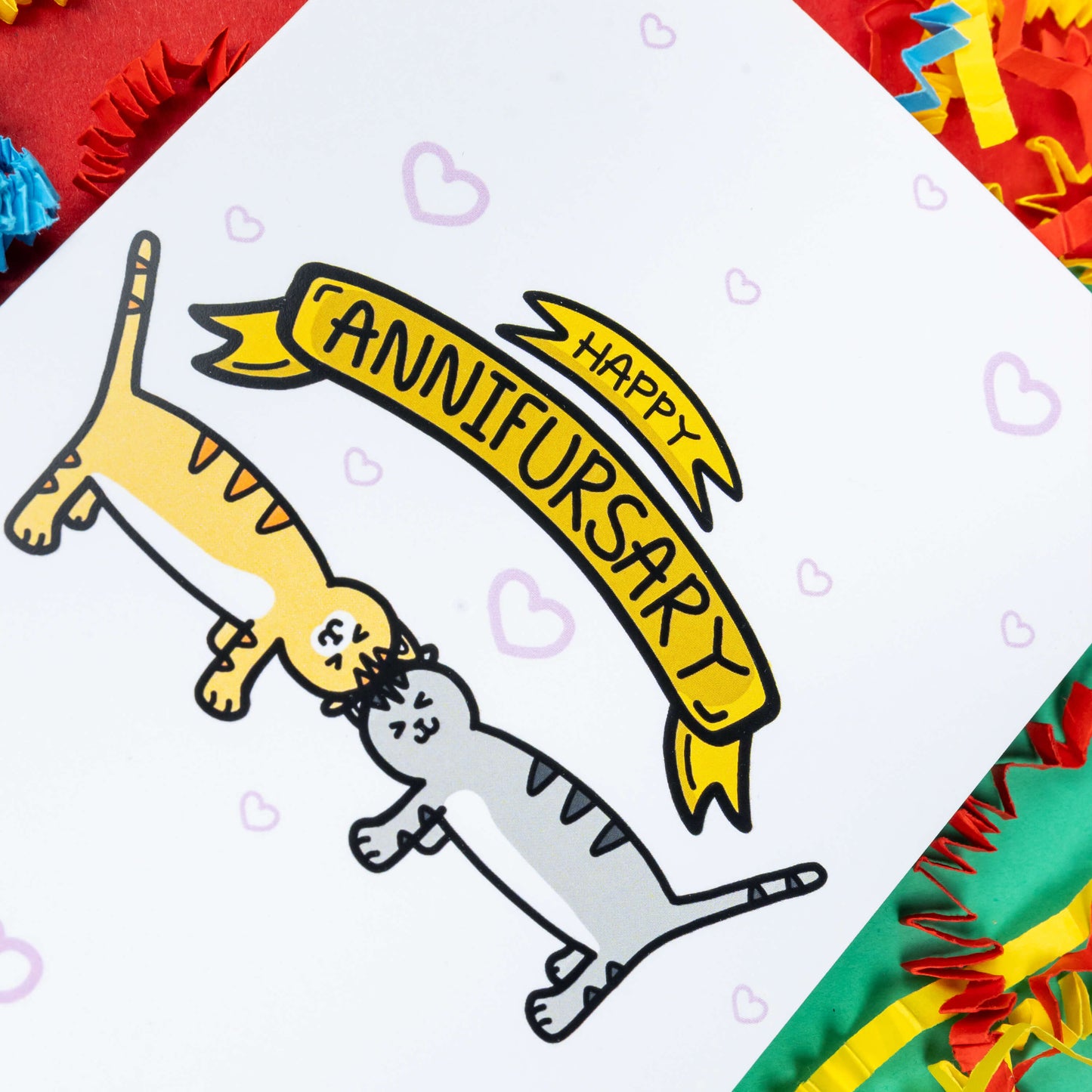a white greeting card with light pink hearts dotted all over. 'Happy ANNIFURSARY' is written in a gold banner on the card with two cat illustrations underneath, one ginger and one grey. The cats are rubbing their heads together. The background of the photo is red, blue and green card with paper card decorations.