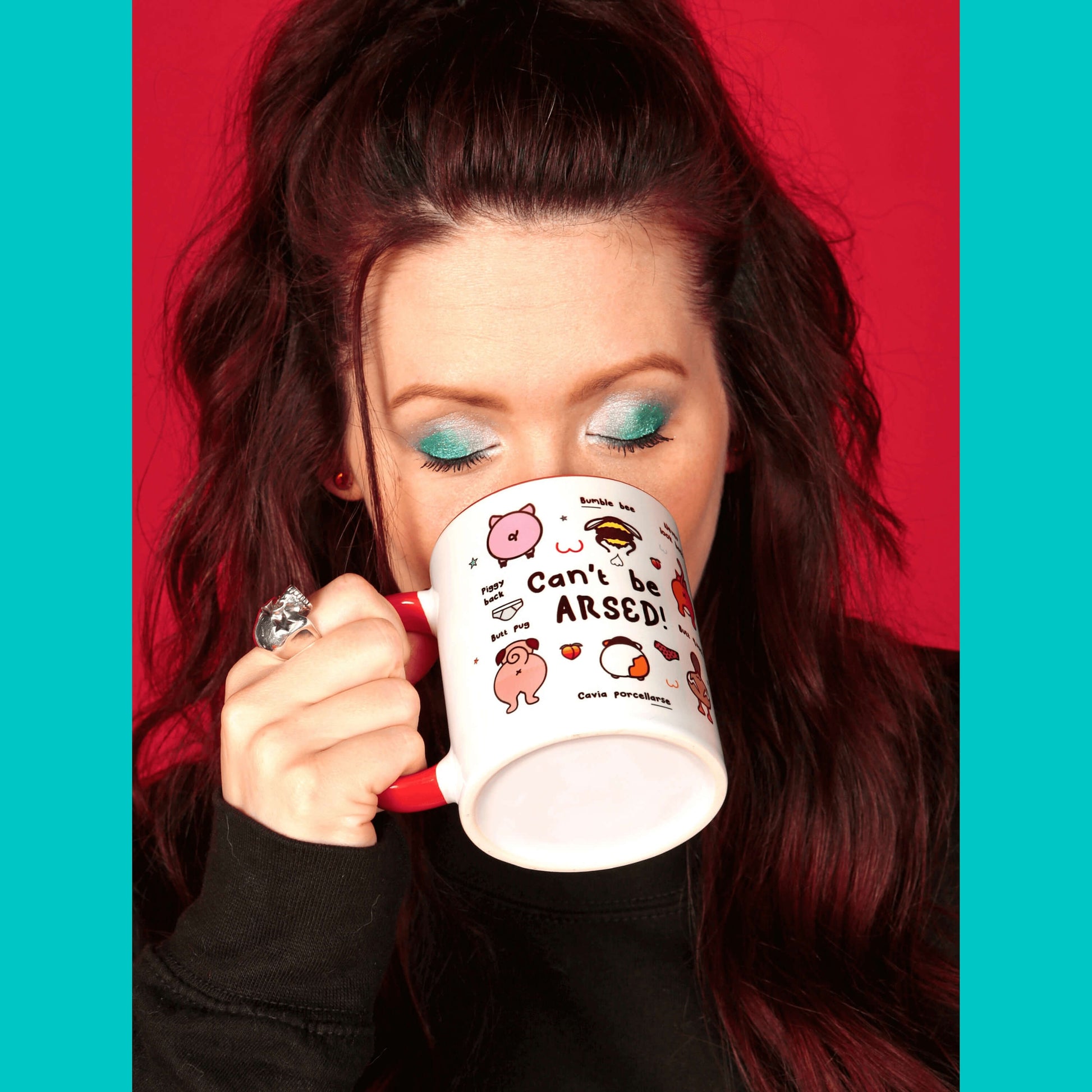 The Can't Be Arsed Mug being held up by Nikky Box to her mouth. She has brown hair and blue eye makeup wearing the invisible illness black sweater. The white mug with a red handle and inside features various animal bums with underwear, chest outlines, peaches and multicoloured stars. The mug is facing right which shows pig - piggy back, bumble bee - bum ble bee, pug - butt pug, guinea pig - cavia porcellarse, duck - butt quack and cat - never look back, bums.