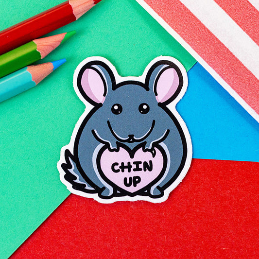 The Chin Up Chinchilla Sticker on a red, green and blue background with colouring pencils and a red stripe candy bag. The sticker is a crouching grey chinchilla holding up a pastel pink heart with black text reading 'Chin Up'.