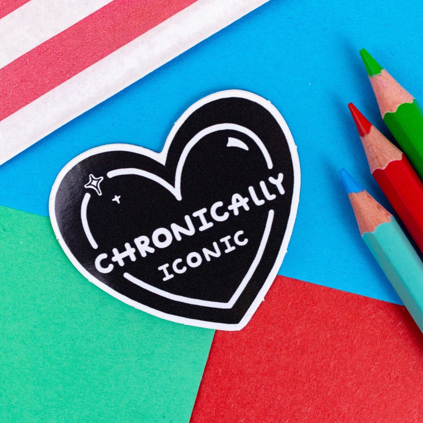 The Chronically Iconic Sticker on a red, blue and green background with colouring pencils and red stripe candy bag. The black heart shaped sticker has a white outline with sparkles and centre text reading 'chronically iconic'. The design is raising awareness for chronic illness and invisible illness.